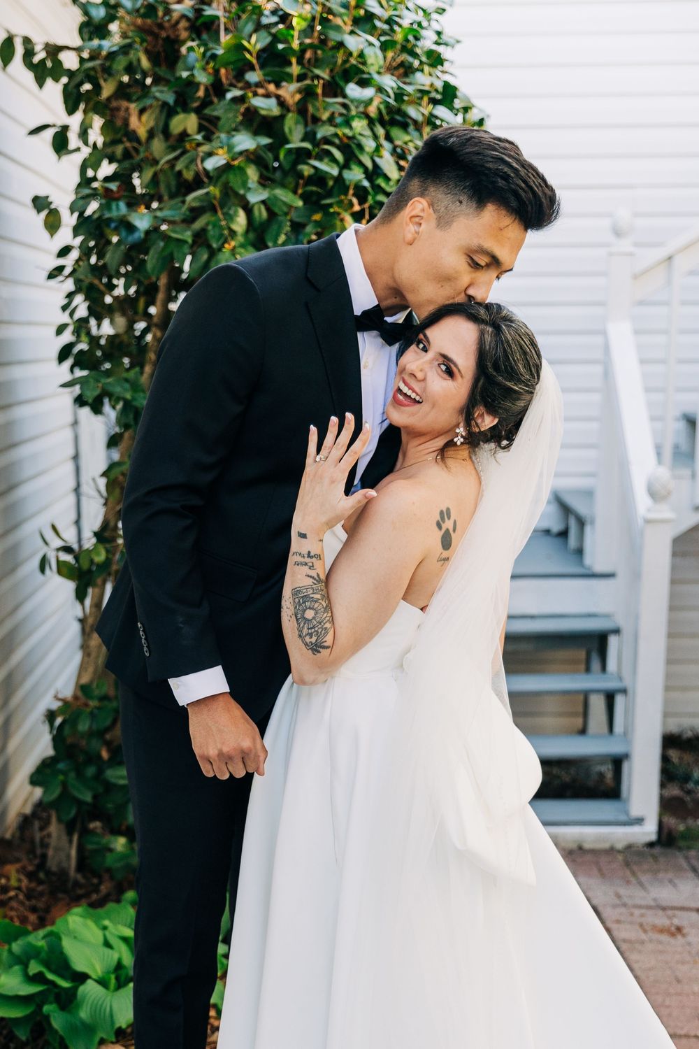 A bride and groom embrace outdoors, she shows her ring and smiles, wearing a white gown, he in a black tuxedo.