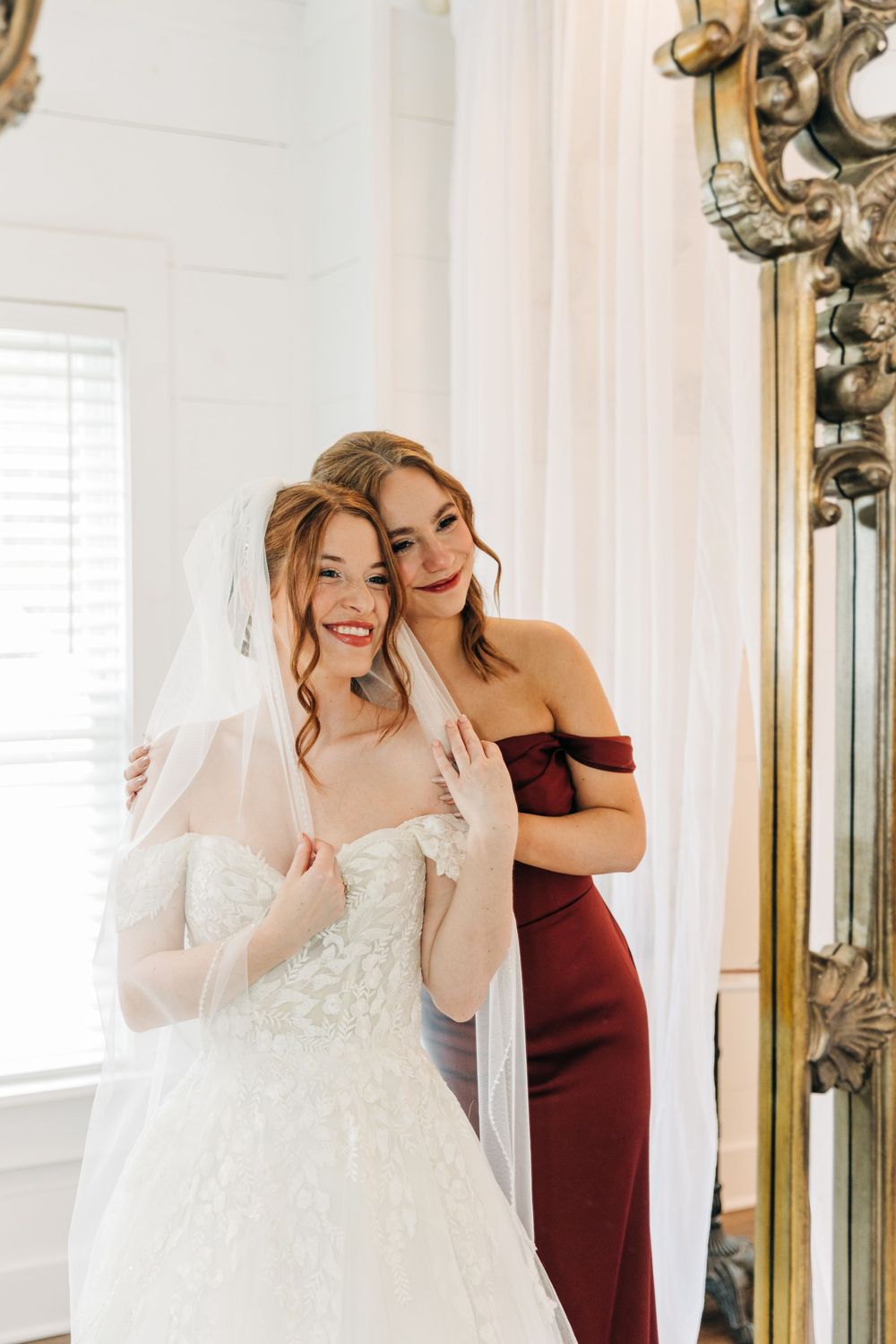 Bride and maid of honor pose together in front of an ornate mirror, one wearing a white wedding gown with veil at Clisby Pines in Georgia