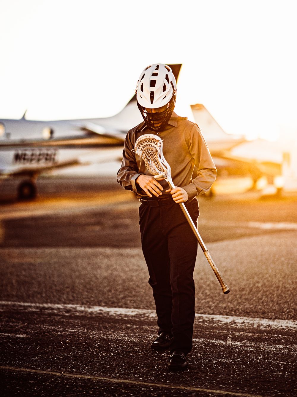 Athlete in protective helmet holding lacrosse stick at sunset near airplane.