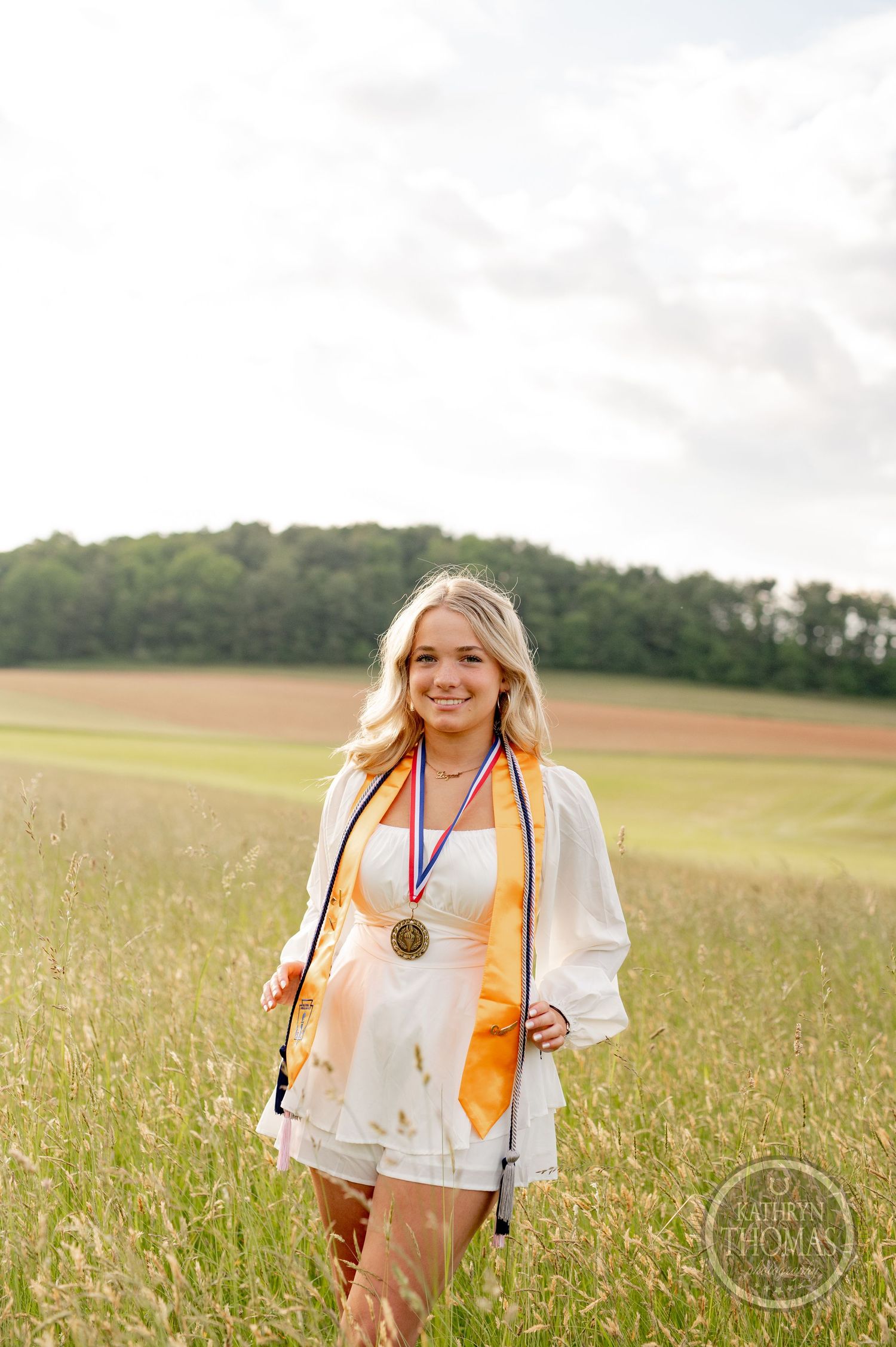 Graduate wearing gold stole and medals stands in sunlit meadow.