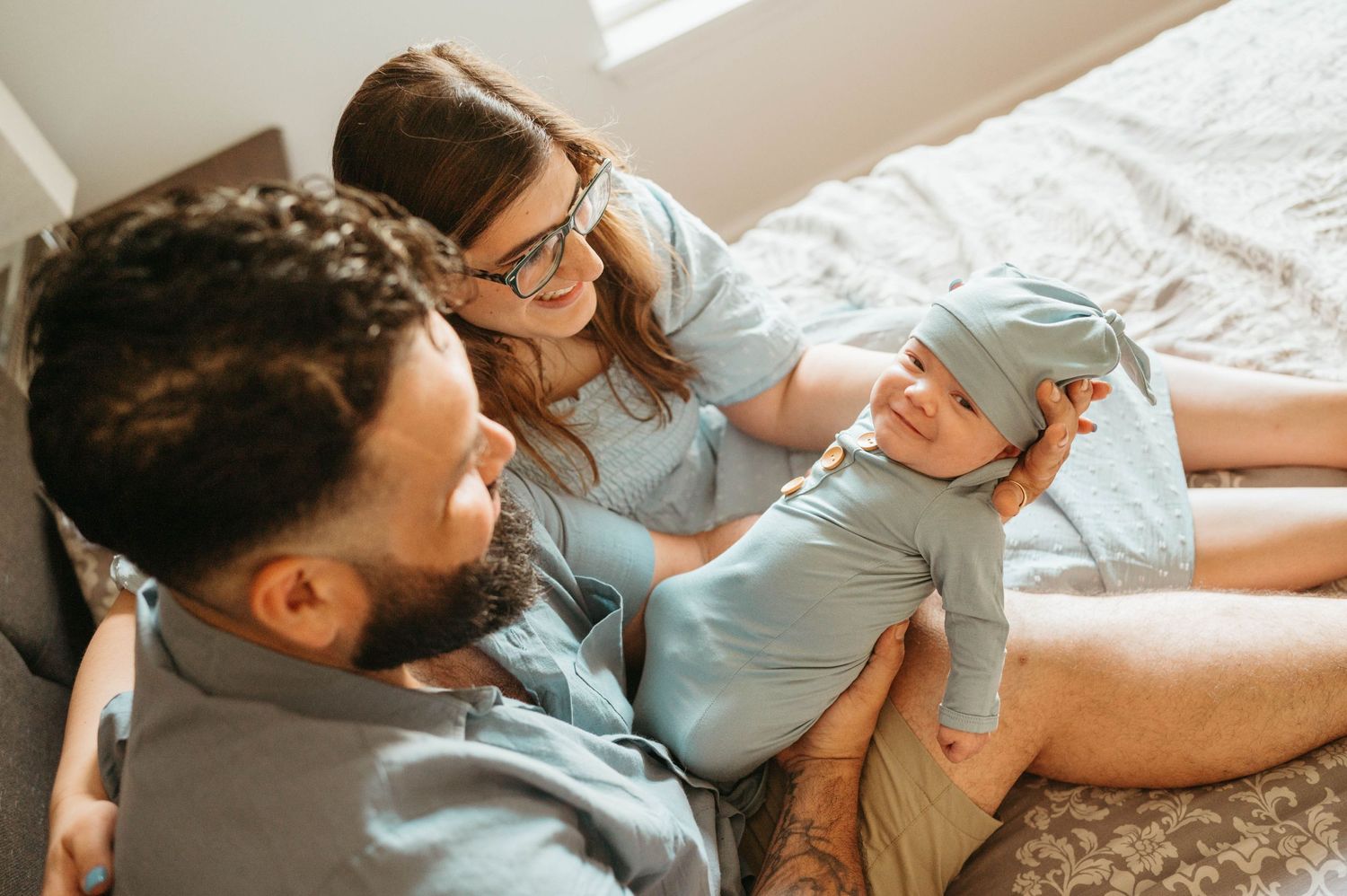 New parents in blue outfits cuddle their alert newborn baby on a bed from an overhead angle.