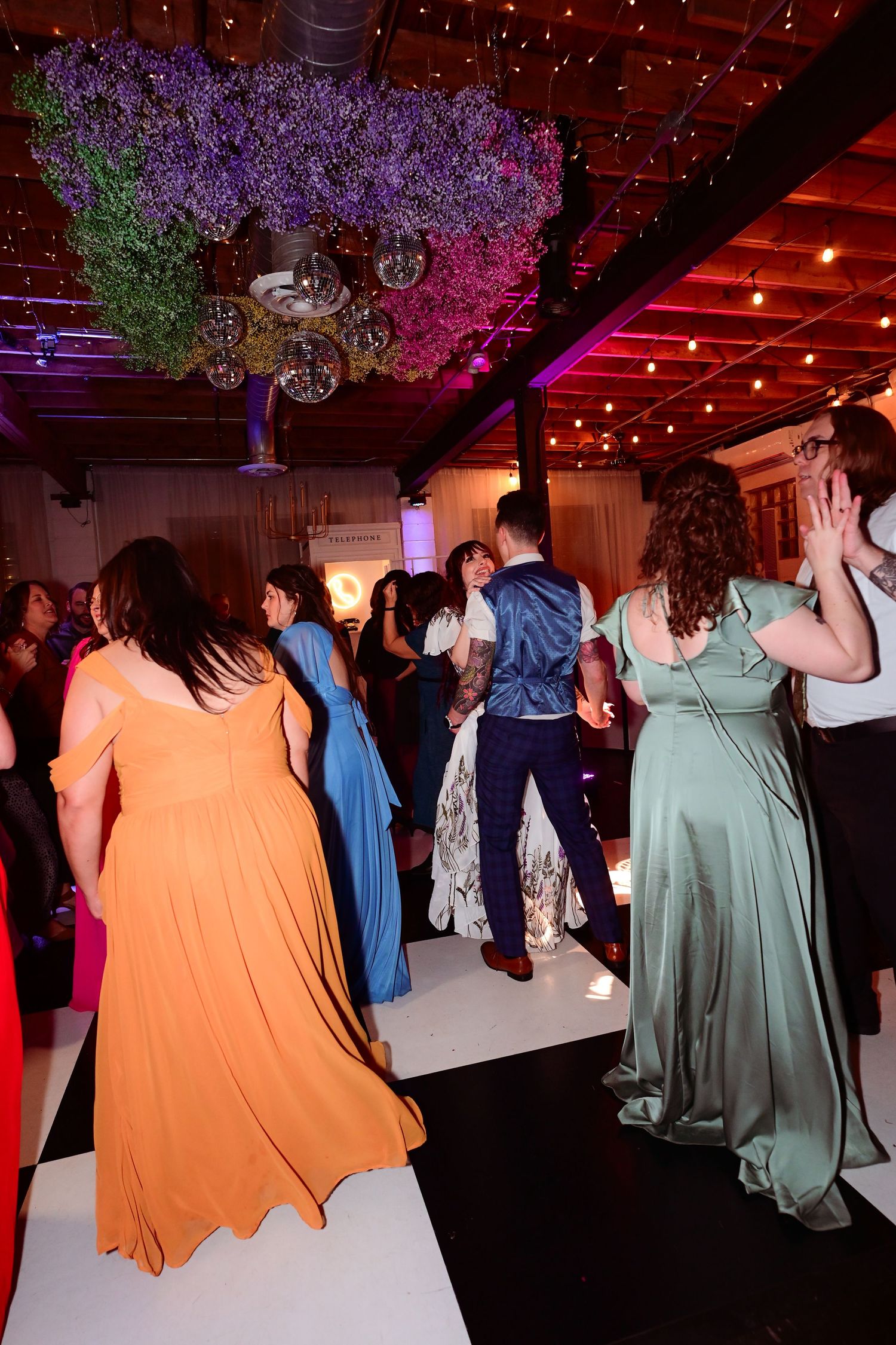 Guests dancing at a wedding reception in formal gowns under floral ceiling decorations and warm string lights.