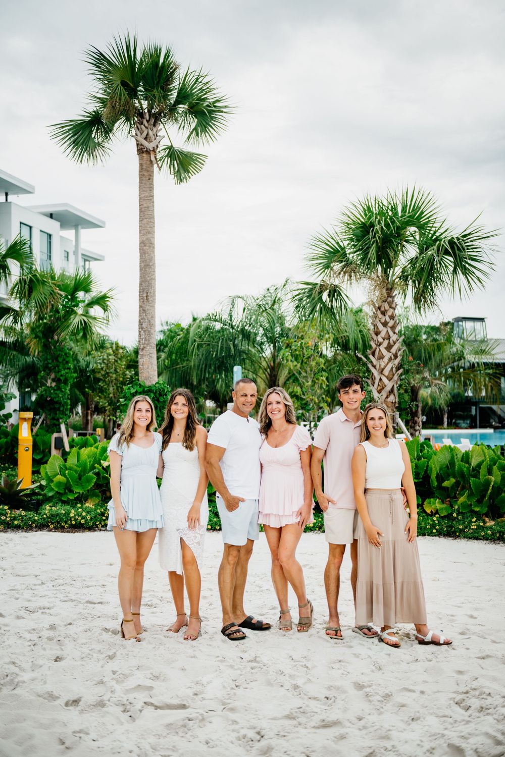 Group in white and neutral outfits poses together on sandy beach with palm trees.