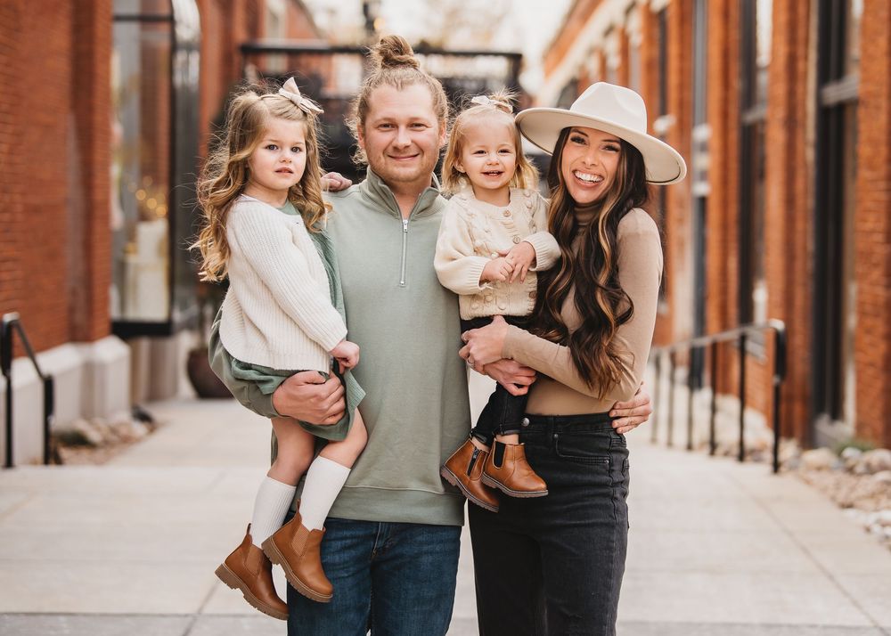 Happy family of four posing on a brick-lined sidewalk, parents holding two young daughters in coordinated fall outfits.