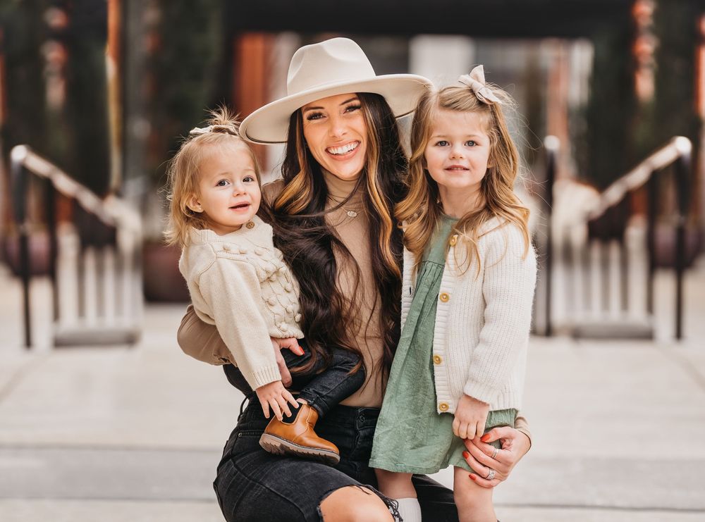 Smiling woman in beige hat holds two young girls outdoors, all dressed in neutral tones for a family photo.