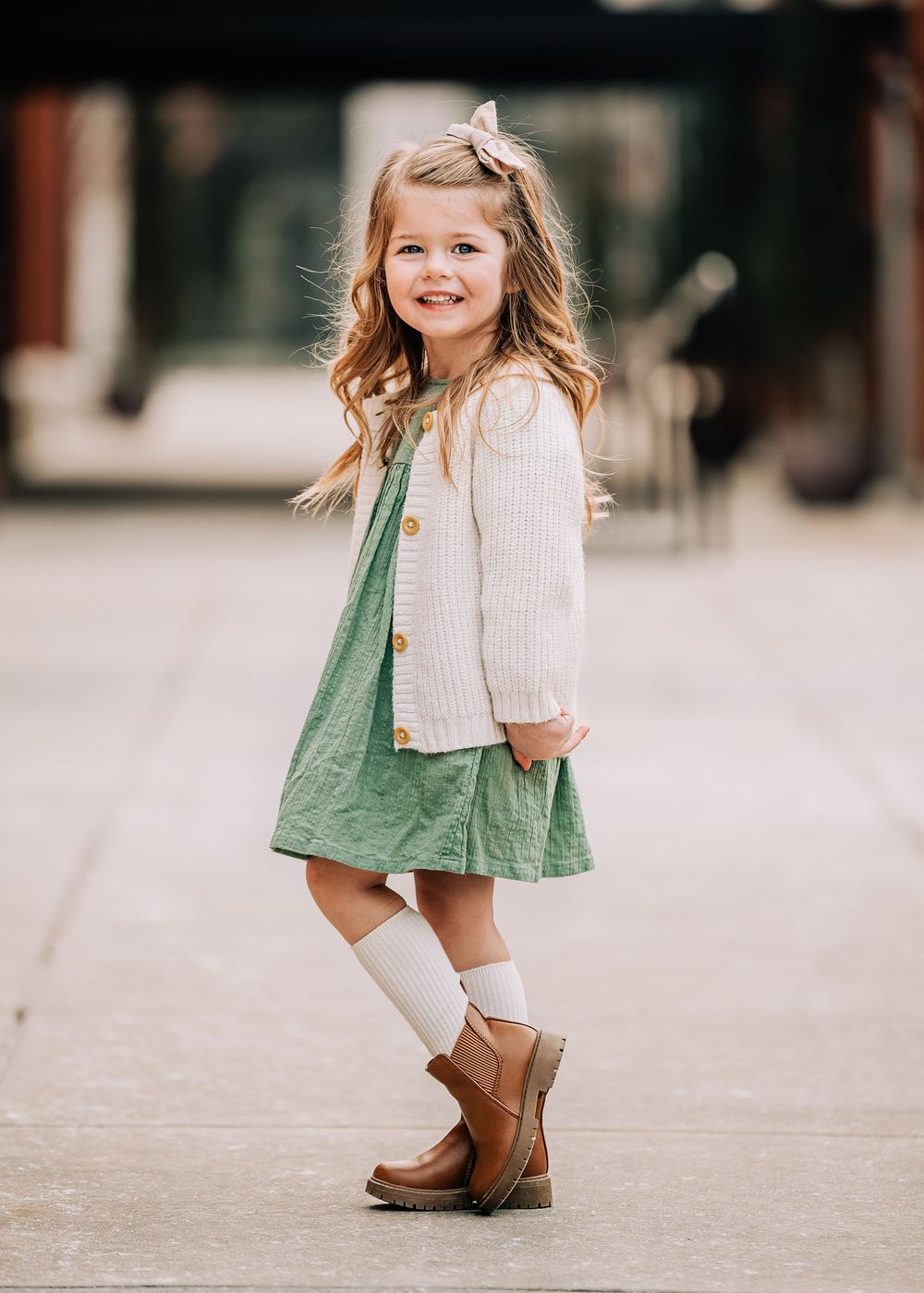 Smiling toddler girl in green dress, cream cardigan, white knee socks, and brown boots with hair bow.