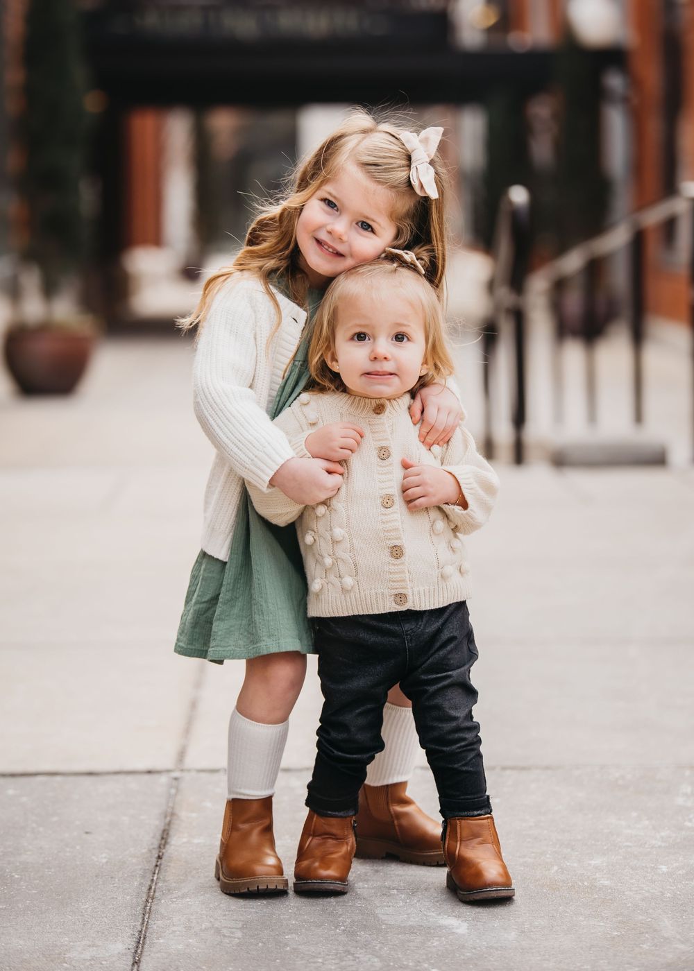 Two young sisters pose together outdoors, older girl hugging younger toddler, both wearing cozy sweaters and brown boots.