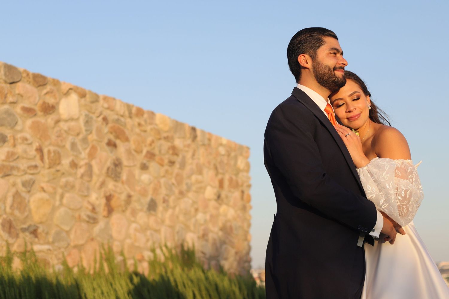 A romantic outdoor portrait of a couple embracing against a stone wall at sunset.