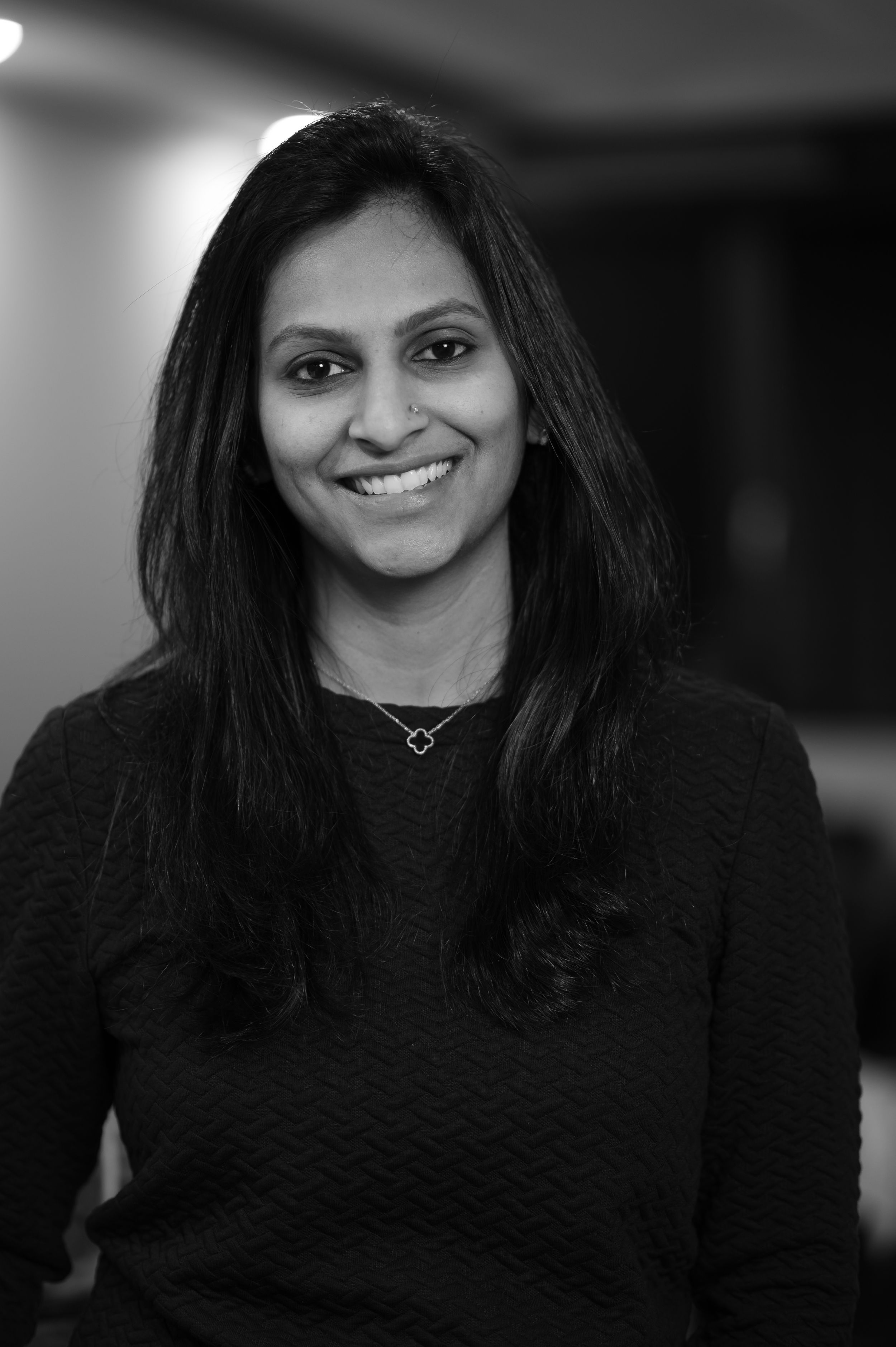 A black and white portrait of a woman with dark hair wearing a dark sweater and necklace, smiling at the camera.