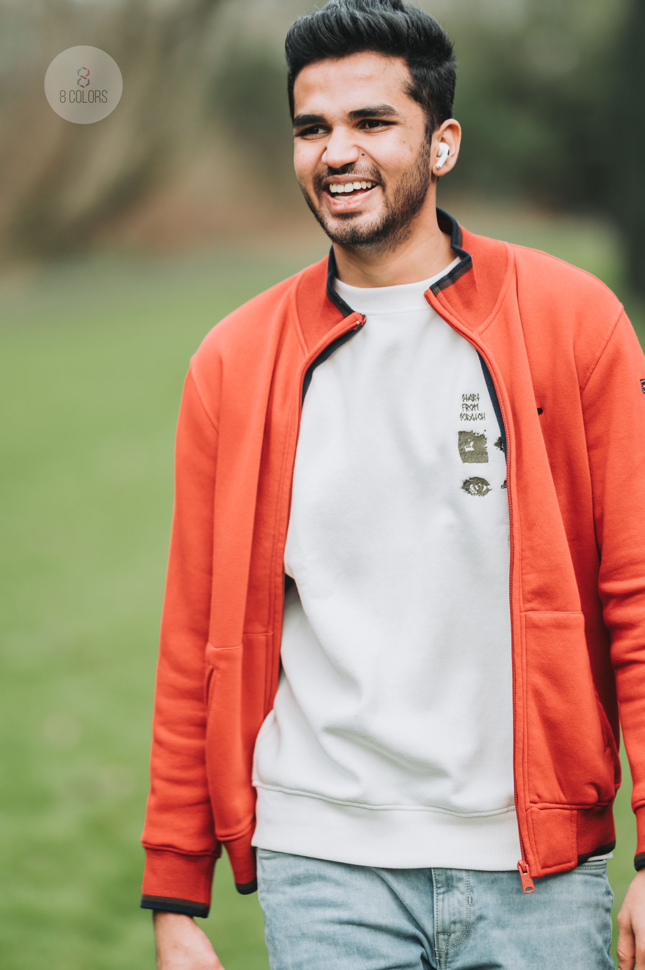 A person in a bright orange jacket and light gray sweatshirt smiles warmly while standing outdoors in a natural setting.