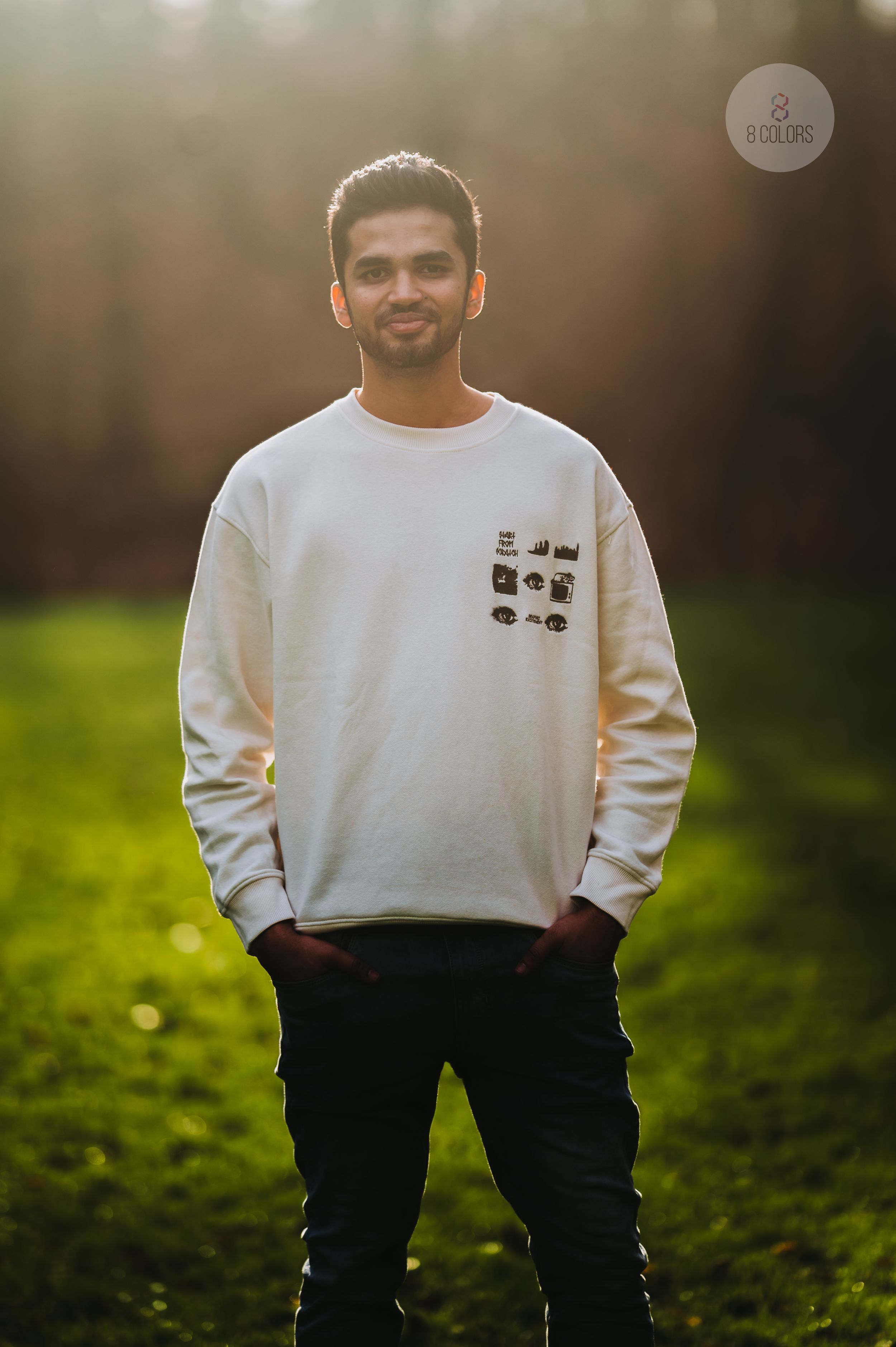 Someone wearing a white crewneck sweatshirt with graphic details and black pants poses in a sunlit grassy area.