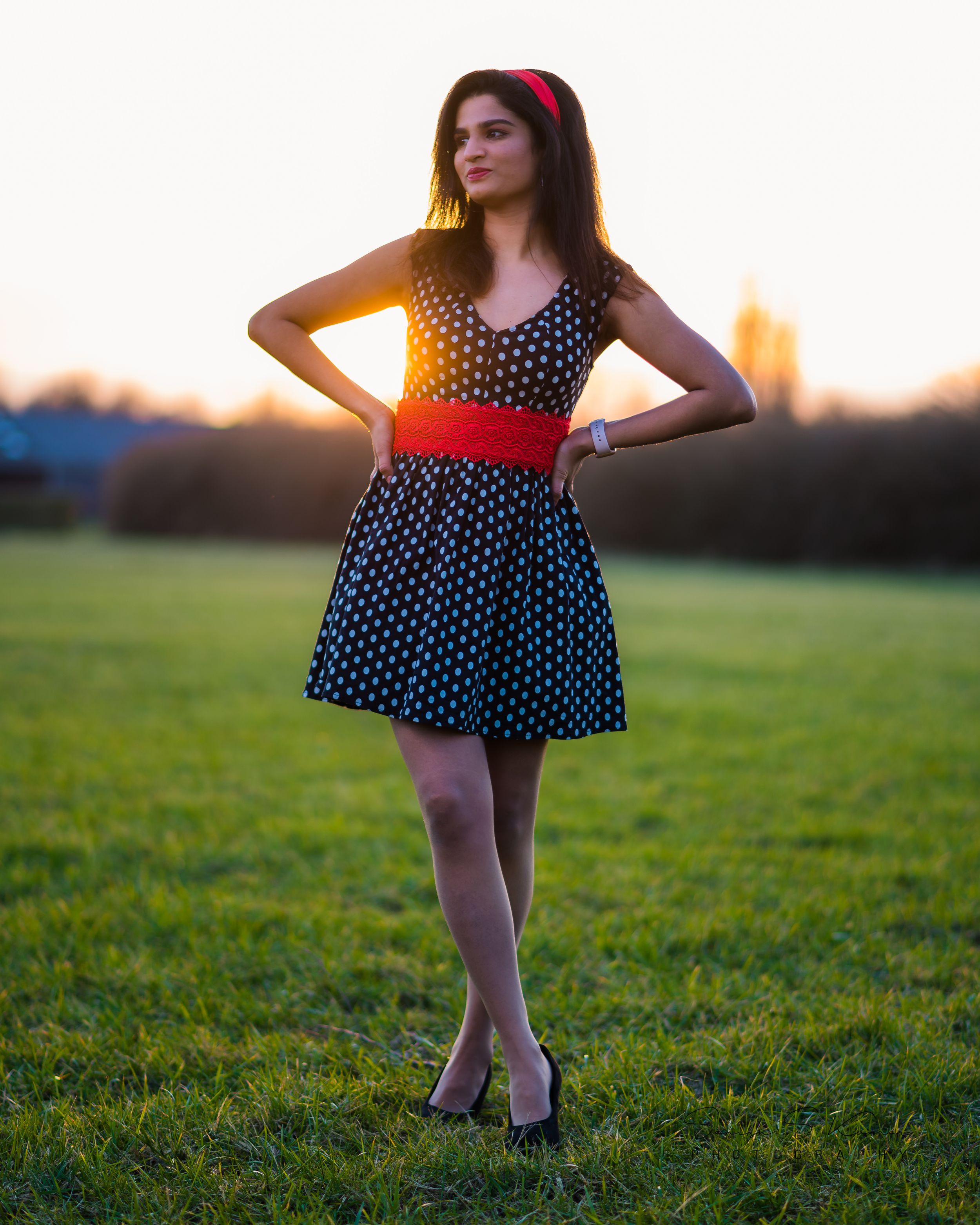 A person in a polka dot dress with red accents poses gracefully on grass during golden hour sunset.