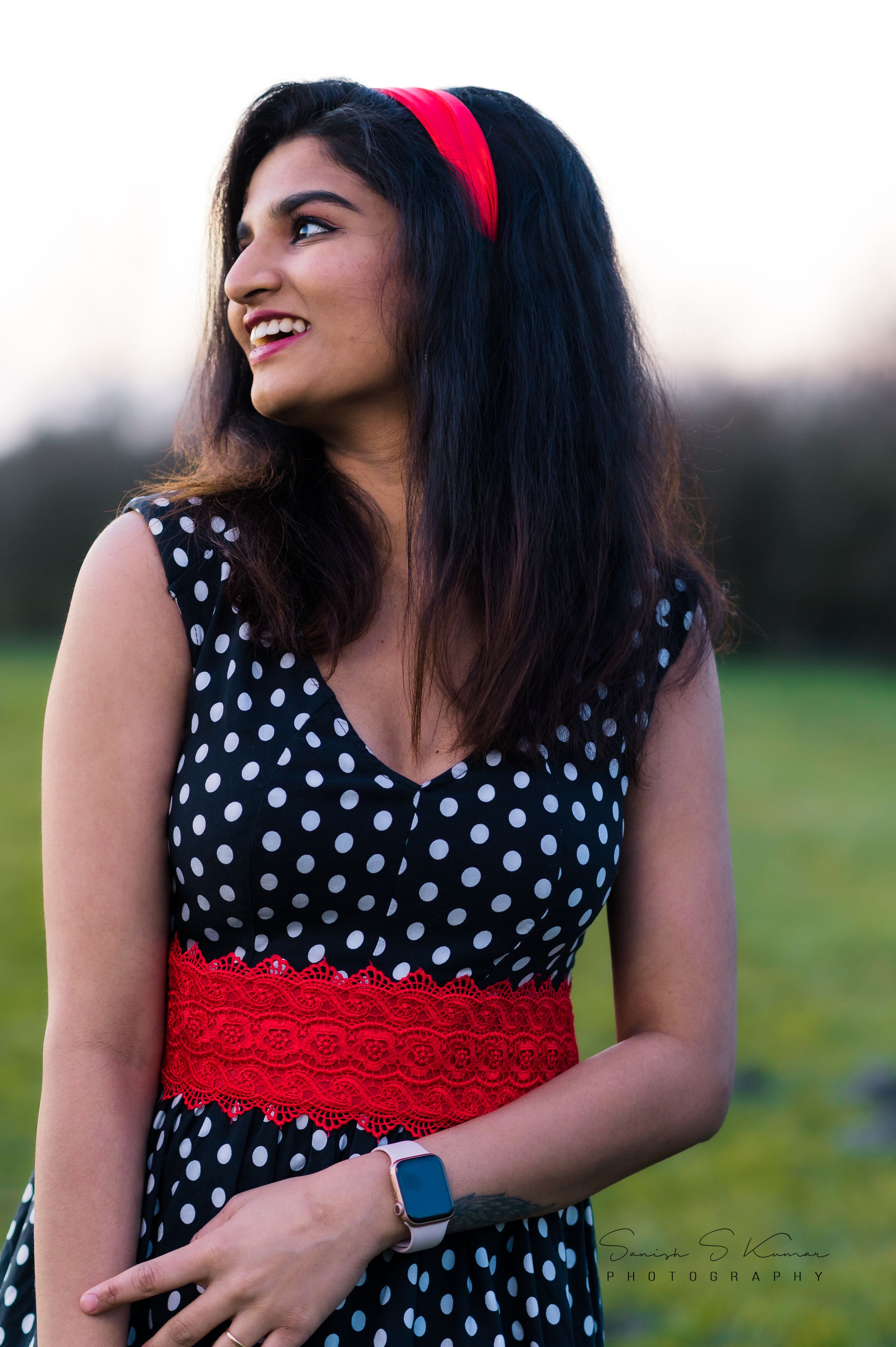 Someone in a polka dot dress and red belt smiles while posing outdoors in the evening light.