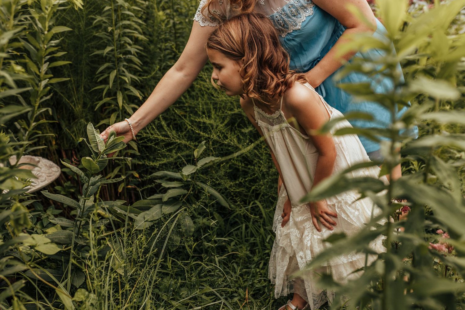 Person wearing a white dress bends down to explore lush green foliage in a summer garden.