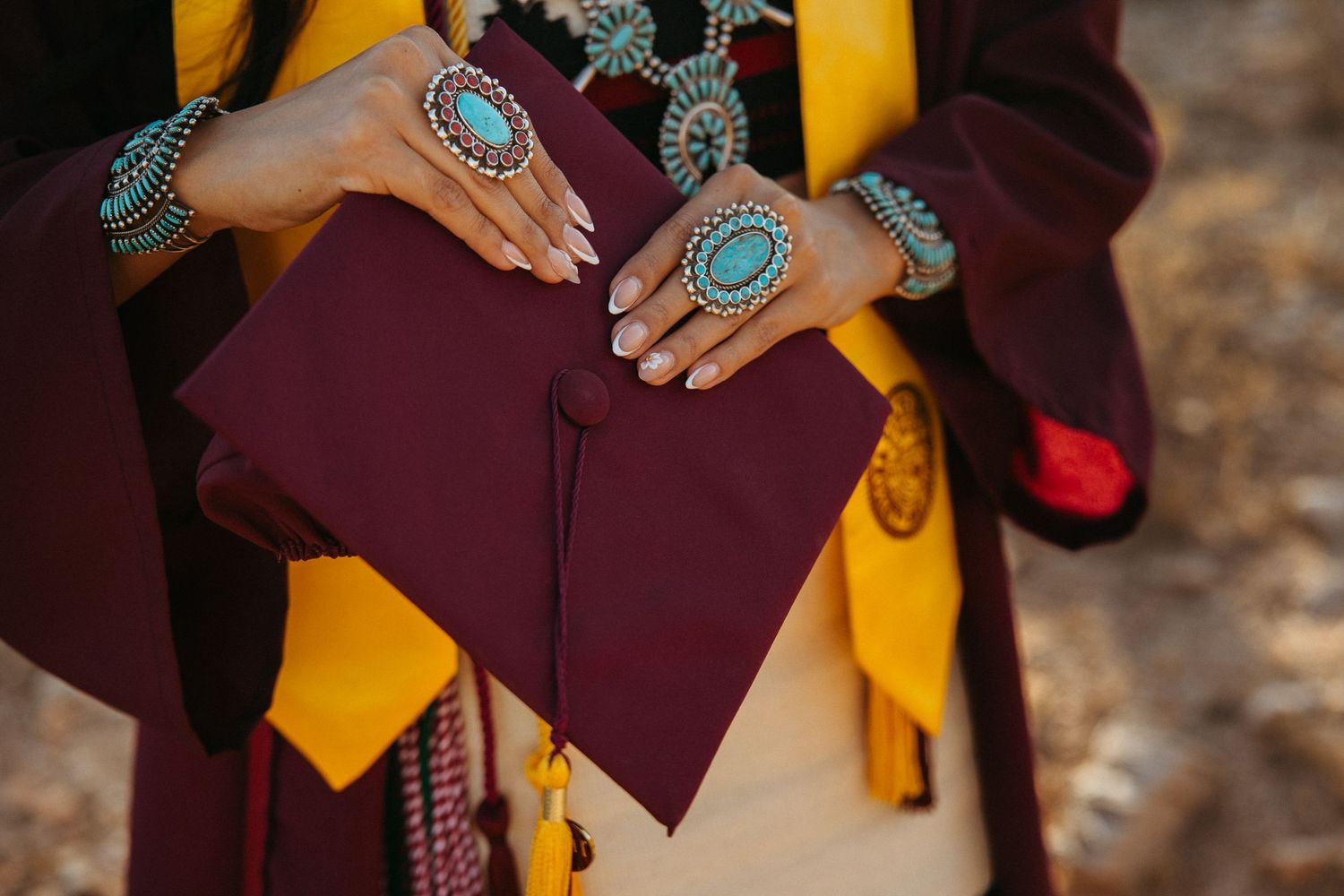 Graduate holding maroon cap adorned with turquoise Native American jewelry, bracelets and rings, wearing yellow and maroon regalia.