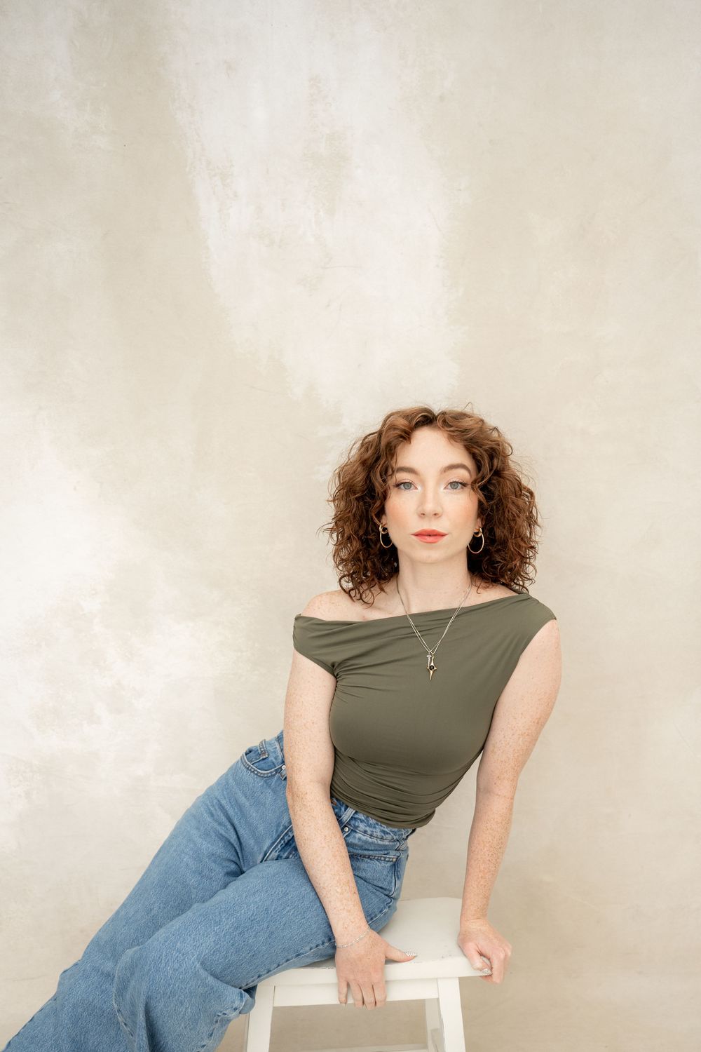 A person in a green off-shoulder top and blue jeans poses casually on a white stool against a neutral backdrop.