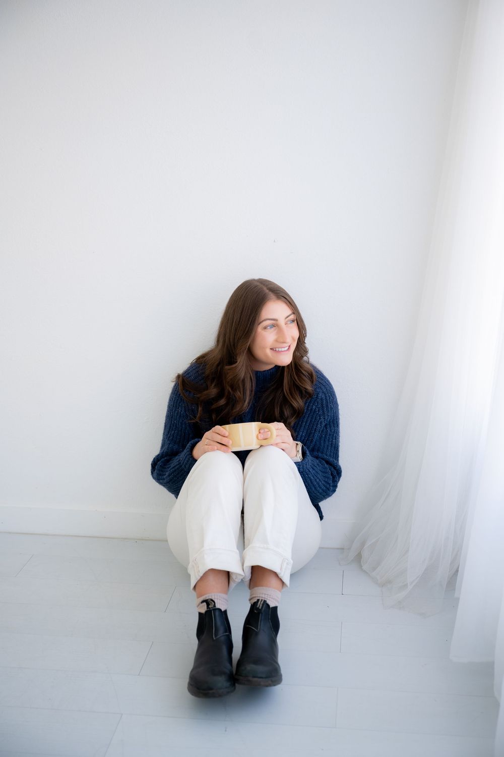 Person in navy sweater and white pants sits casually against white wall wearing black boots.