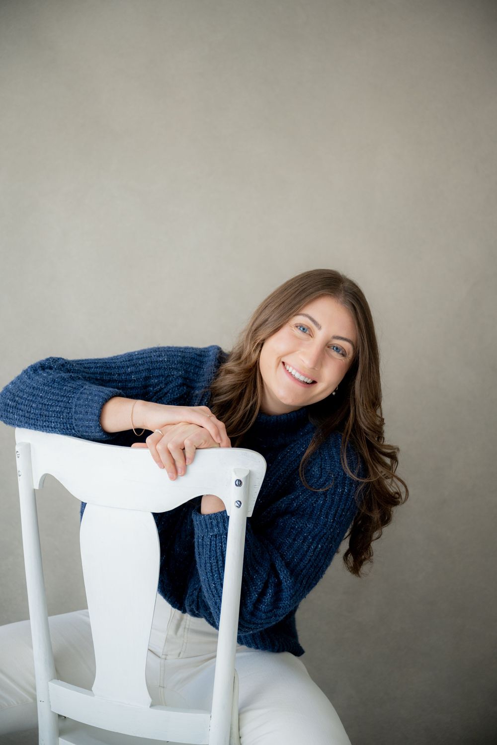 A person wearing a navy blue sweater poses playfully on a white chair against a neutral gray background.