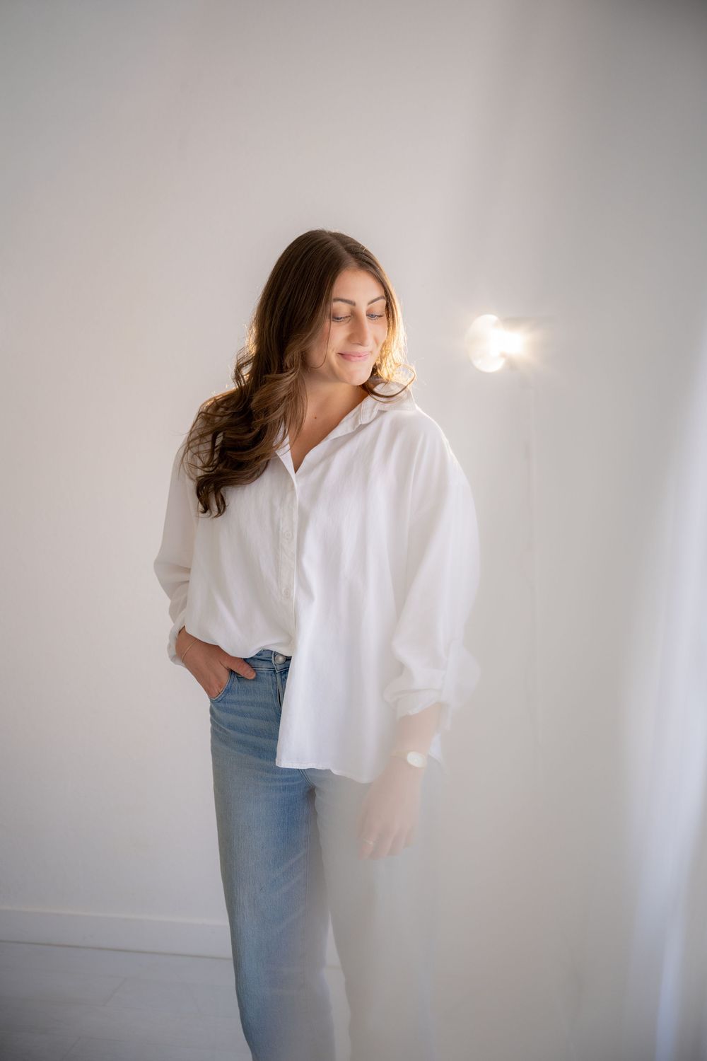 A person in a white button-down shirt and blue jeans poses against a light gray wall in a minimalist style photoshoot.