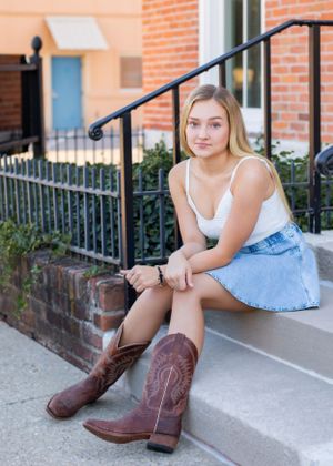 Person in denim skirt and brown cowboy boots sits on concrete steps near brick building.
