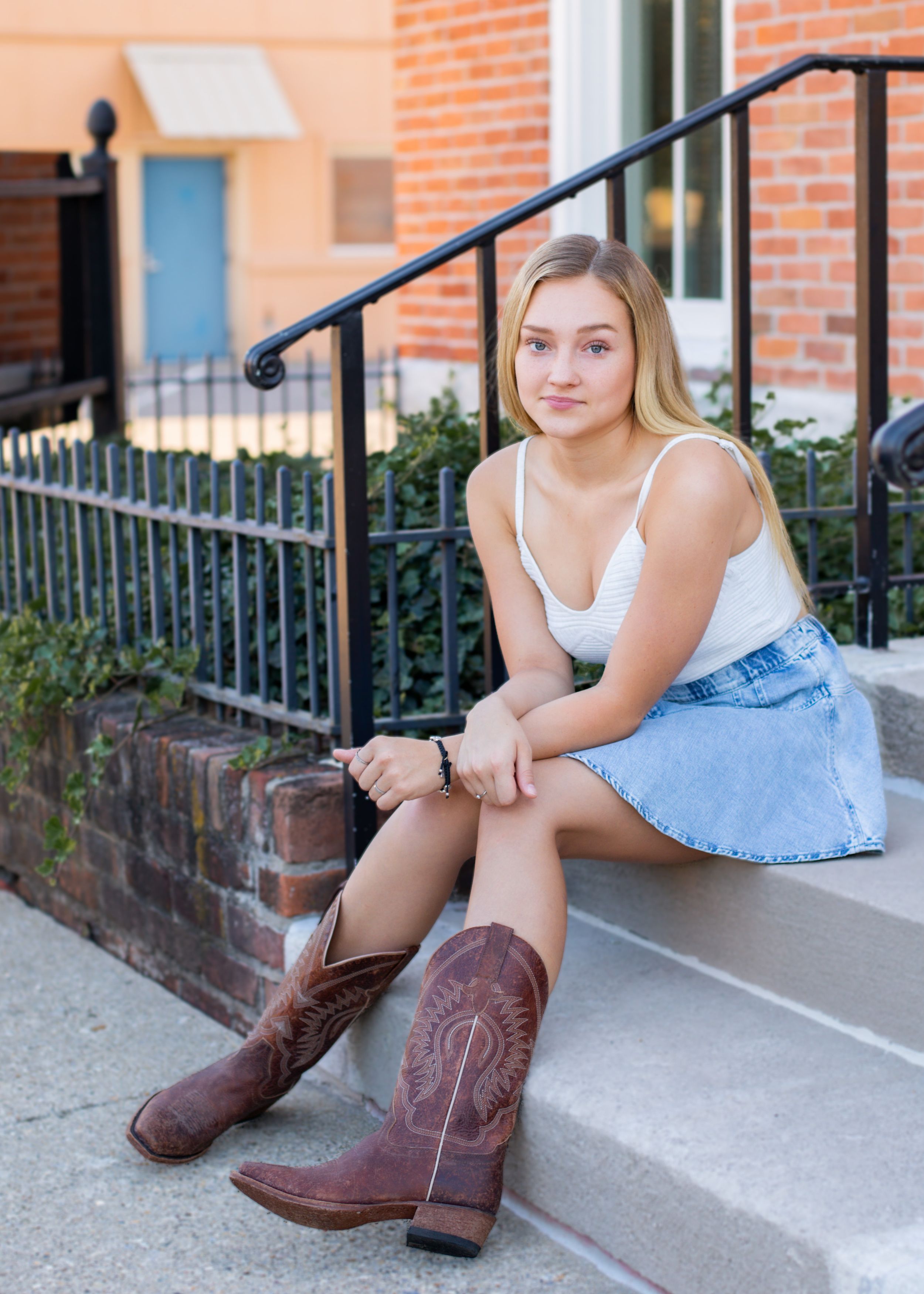 Person in denim skirt and brown cowboy boots sits on concrete steps near brick building.