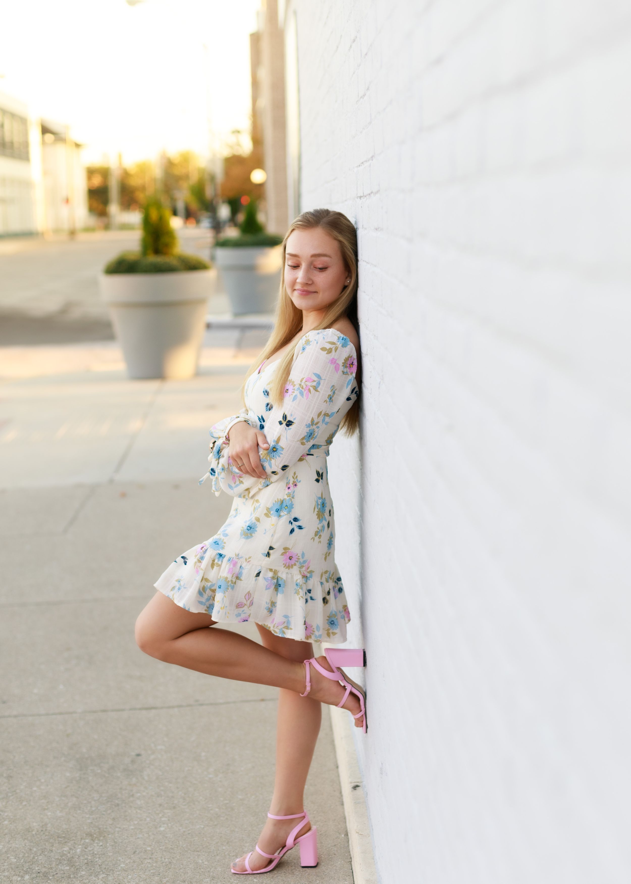 Summer fashion poses in a floral dress and pink heels against a white building exterior.