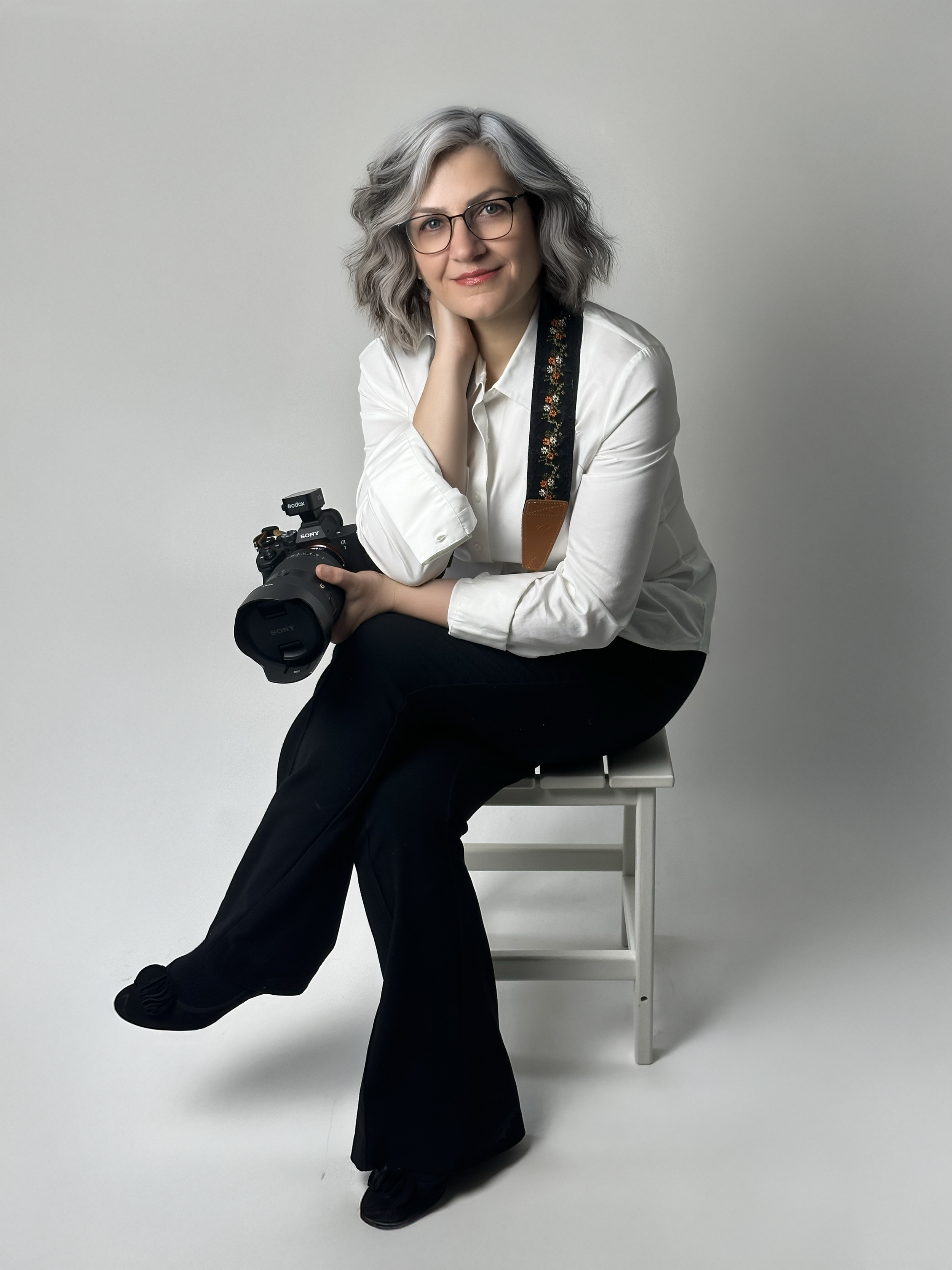 A woman with gray hair and glasses sits on a white stool holding a camera, wearing a white blouse and black flare pants.
