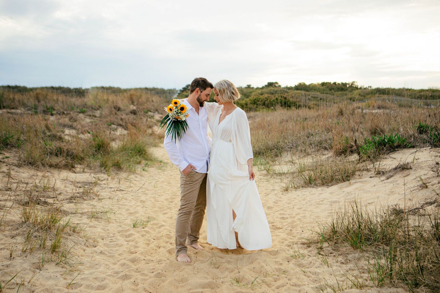 Intimate beach engagement photoshoot among coastal dunes and vegetation.