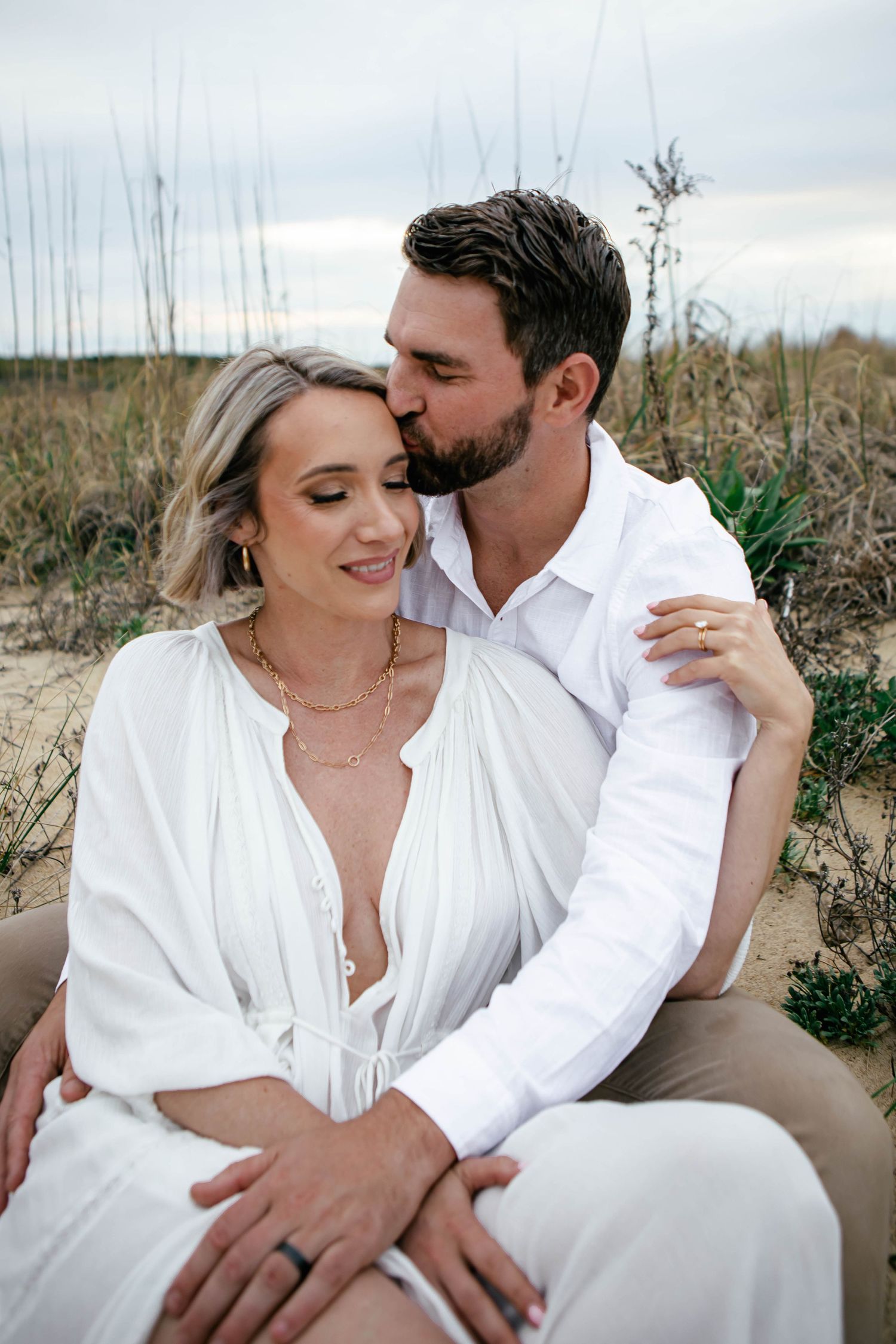 Romantic beach portraits with couple in white clothing sitting on sandy dunes.
