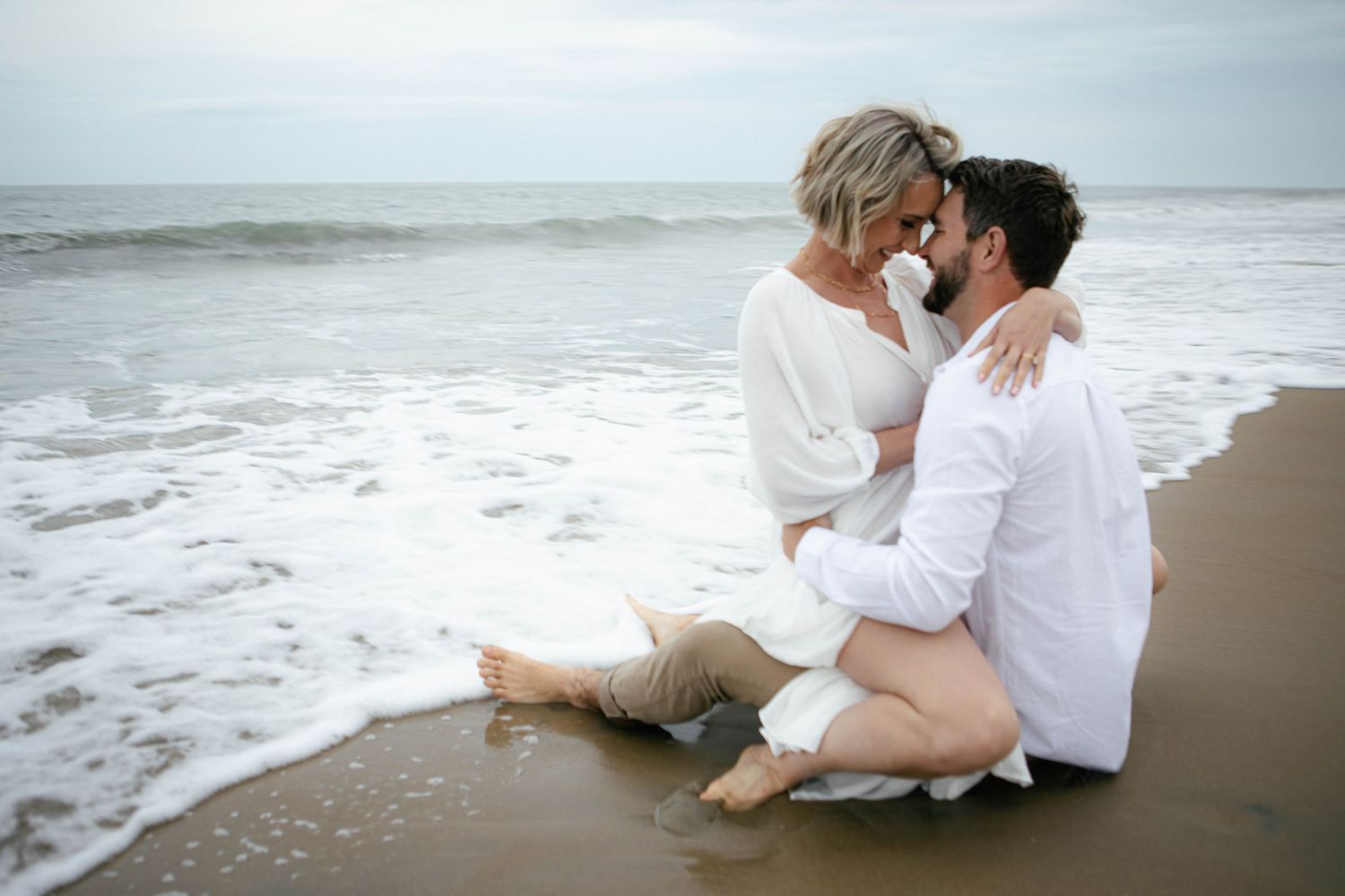 Couple in white clothing sitting together on wet sand at the ocean's edge.