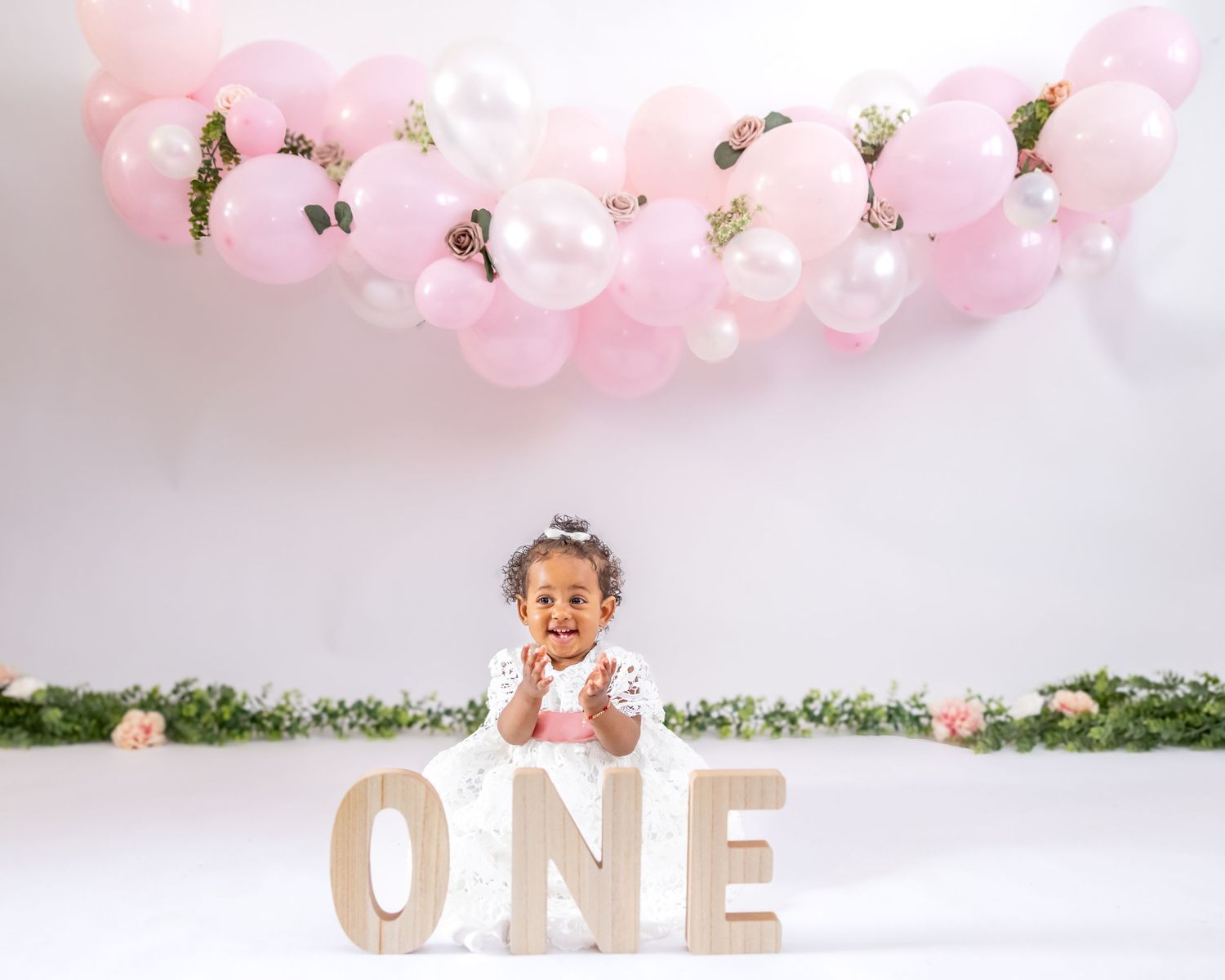 Pink and white balloon garland decorates a first birthday photo setup with wooden 'ONE' letters on white backdrop.