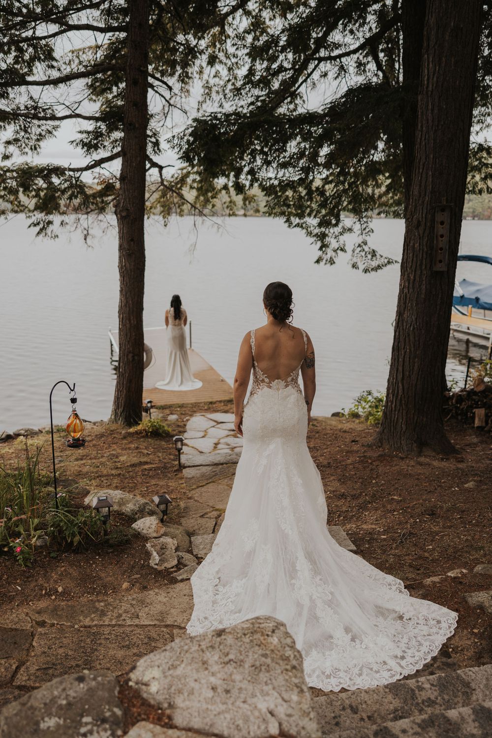 Two wedding dresses with flowing lace trains photographed against a scenic lakeside backdrop with towering pine trees.
