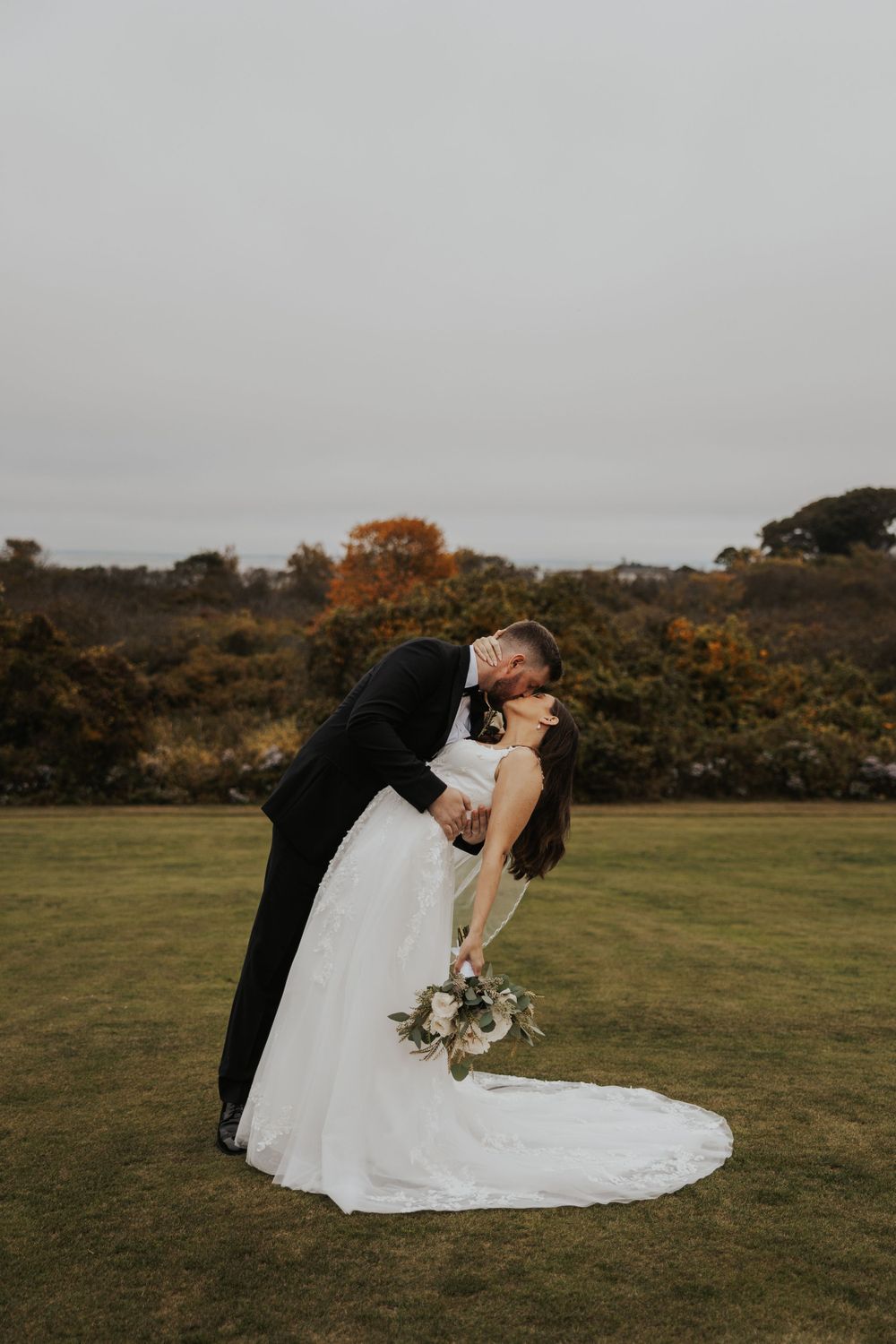 Dramatic wedding portrait with couple sharing romantic dip pose on grassy hillside.