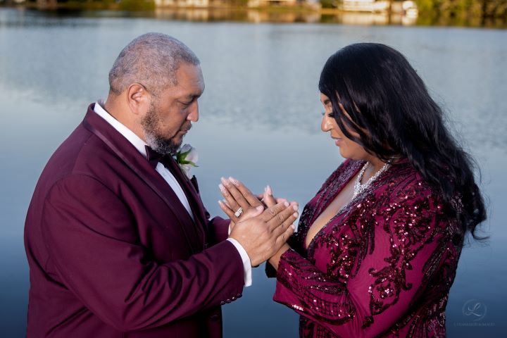 Couple in elegant burgundy attire share an intimate moment by a waterfront setting.