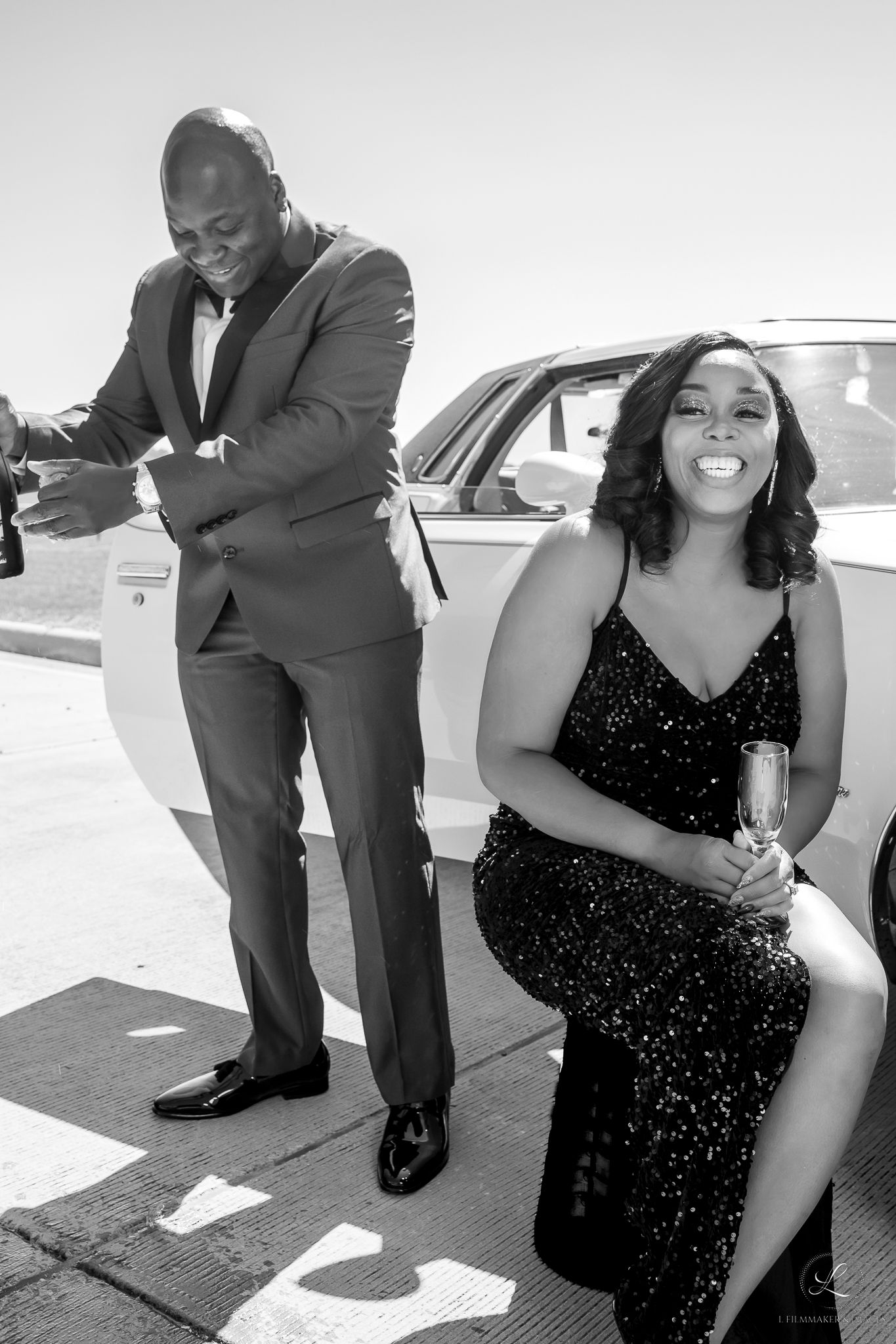 Black and white photo of couple in formalwear posing playfully near a luxury vehicle.