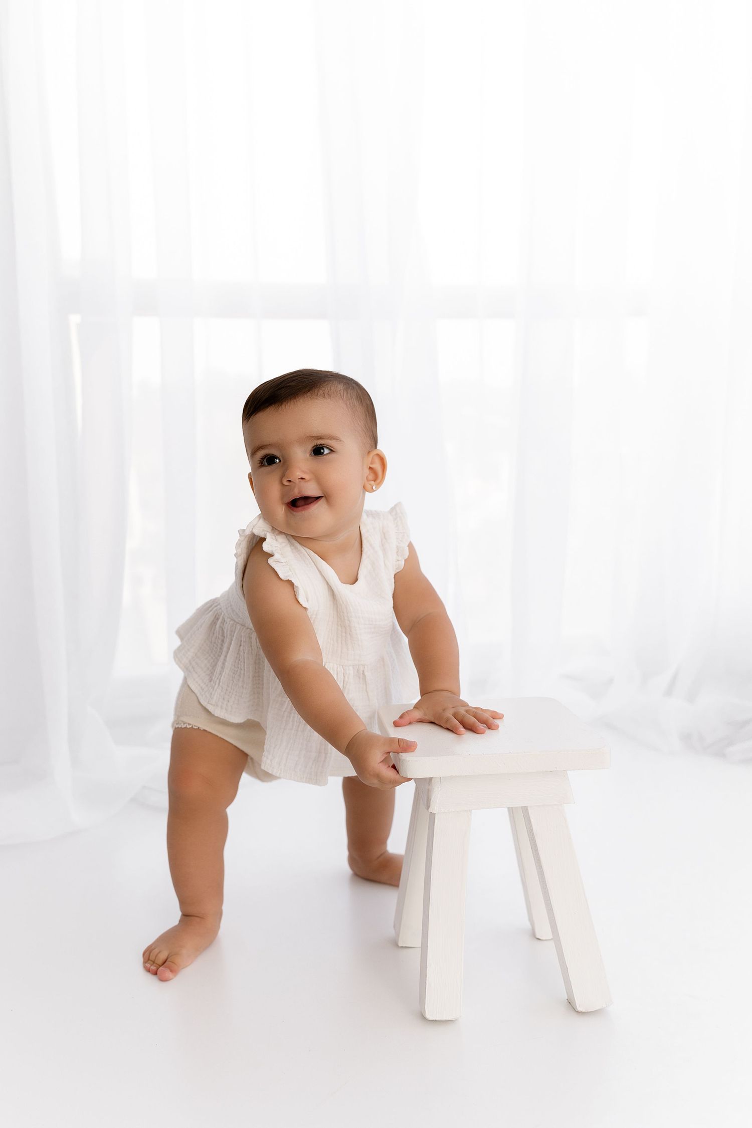 Baby in white outfit practices standing while holding onto small white stool in minimalist setting.