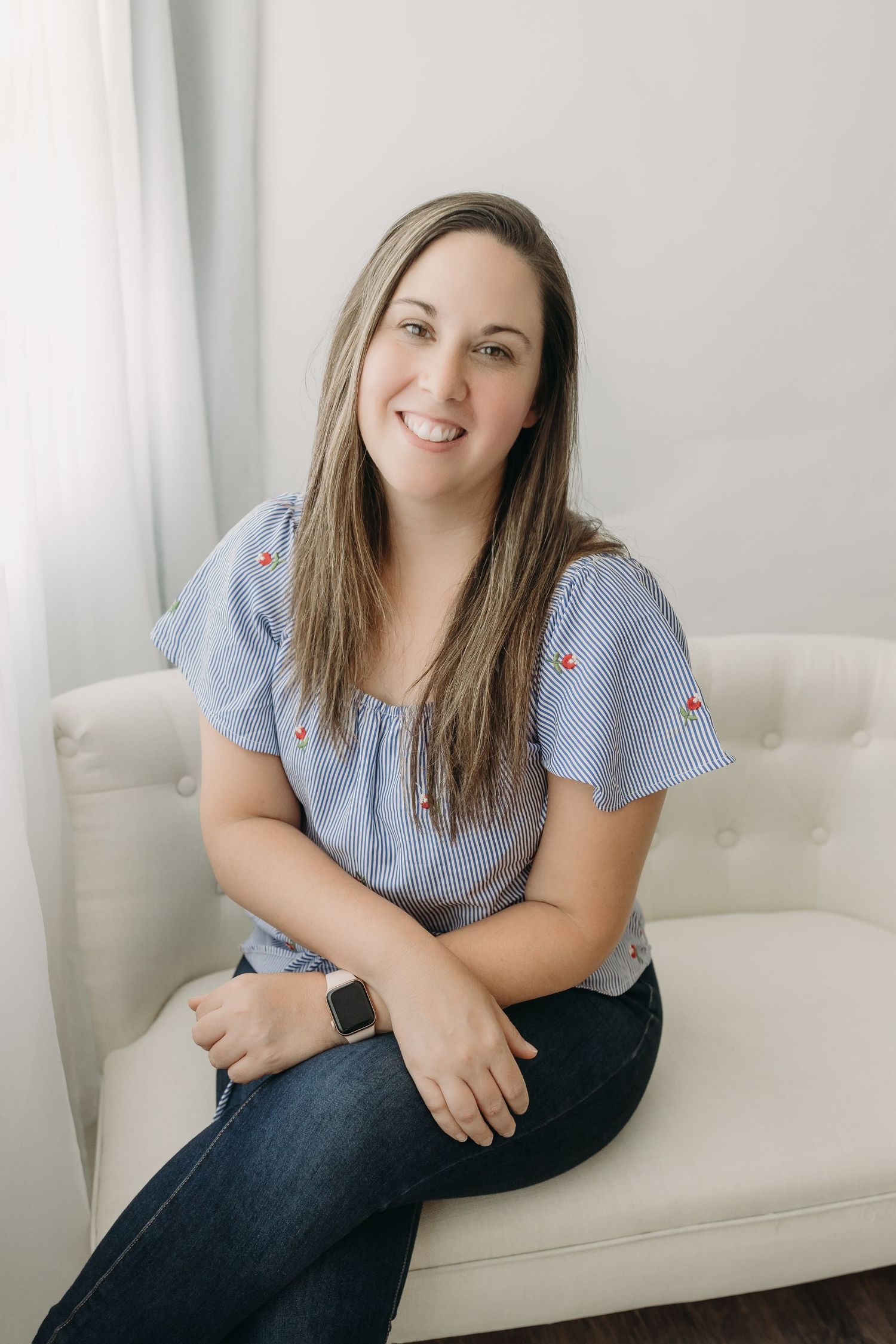 Tampa Photographer in light blue floral top and jeans sits comfortably on white couch against light background, LB Photography, LLC