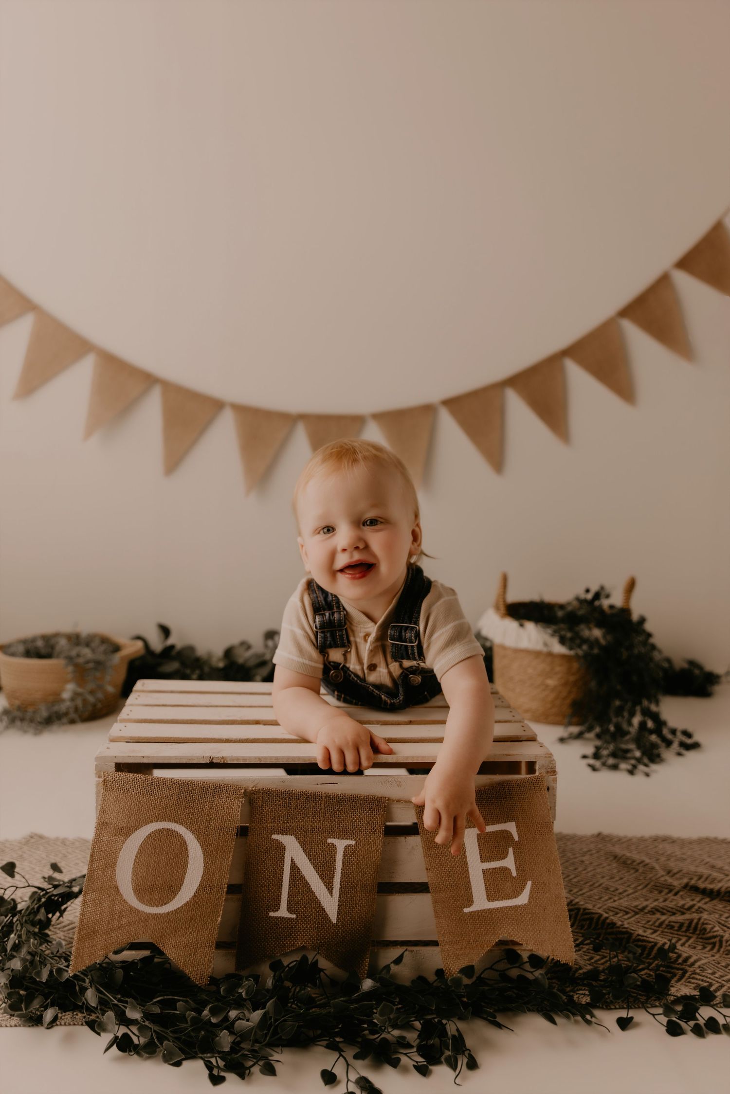 Smiling baby boy celebrating his first birthday sitting in a wooden crate with burlap ONE banner and bunting.