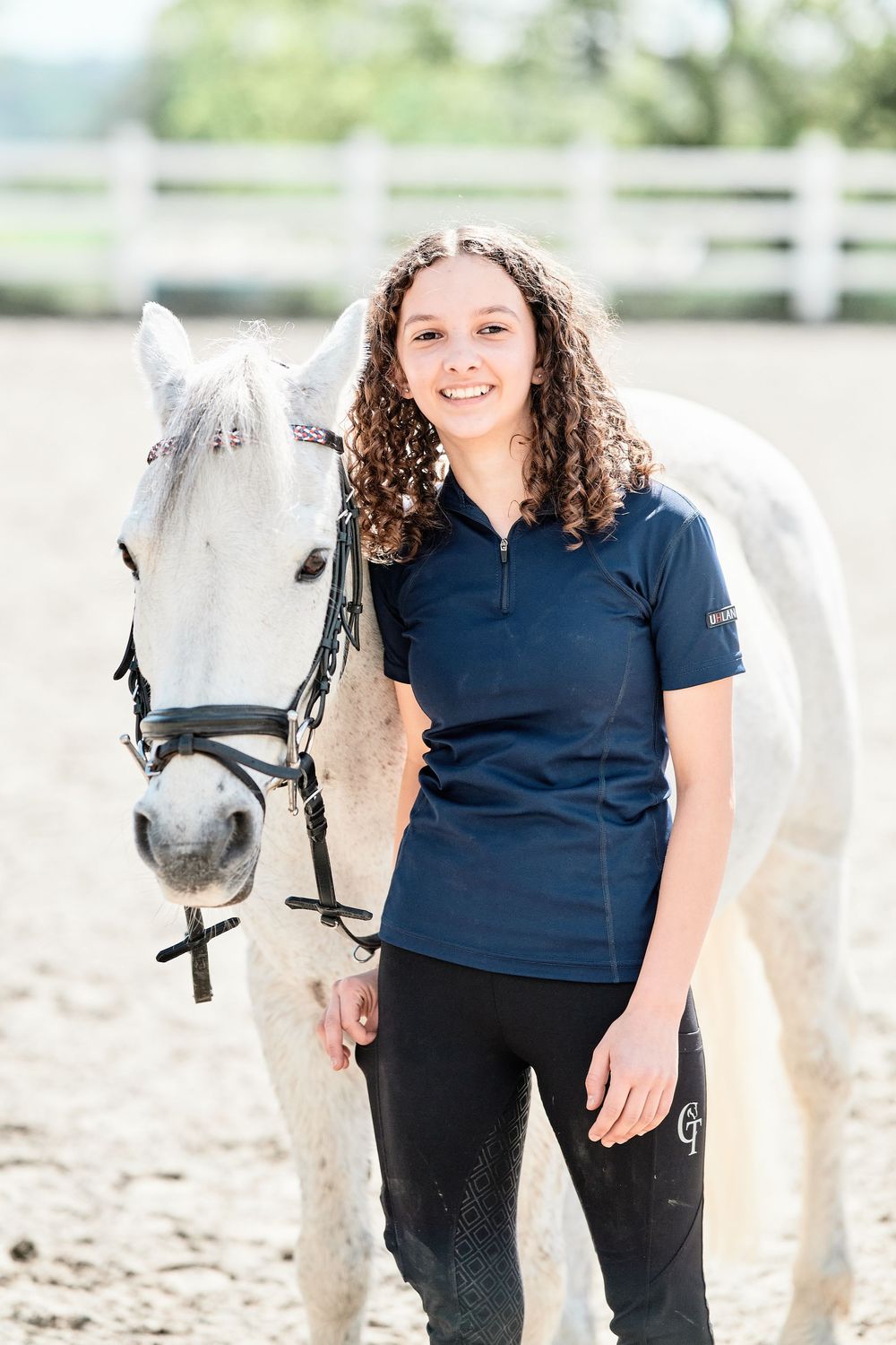 Equestrian stands with beautiful white horse in riding arena wearing navy polo shirt and black riding pants.