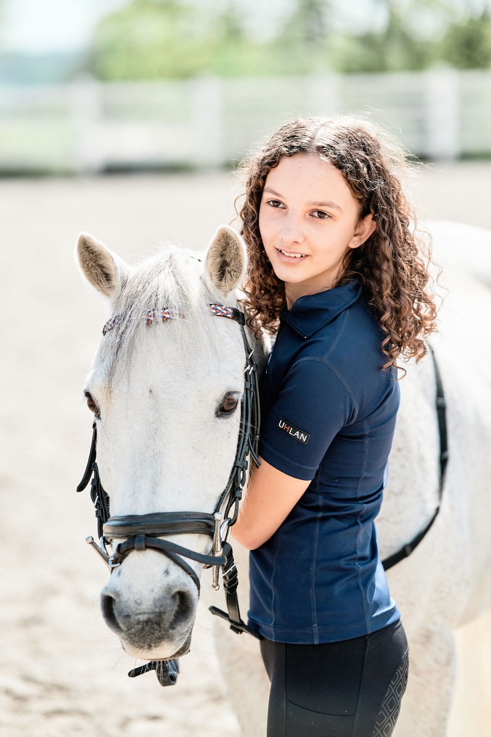 A person with curly hair poses with a friendly white horse wearing a black bridle in an equestrian setting.