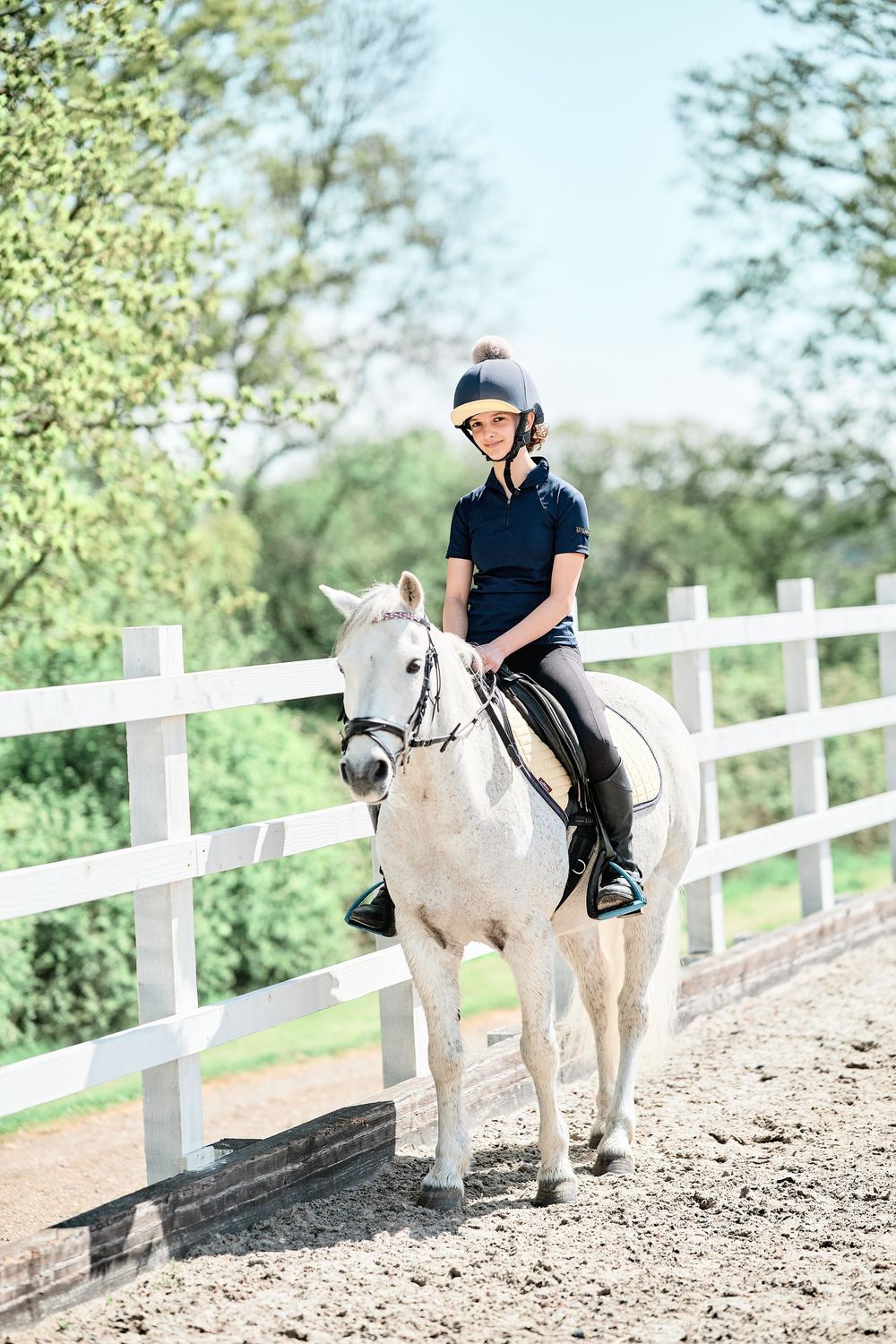 Equestrian rider in navy polo and helmet riding a white horse along a white fenced arena on a sunny day.