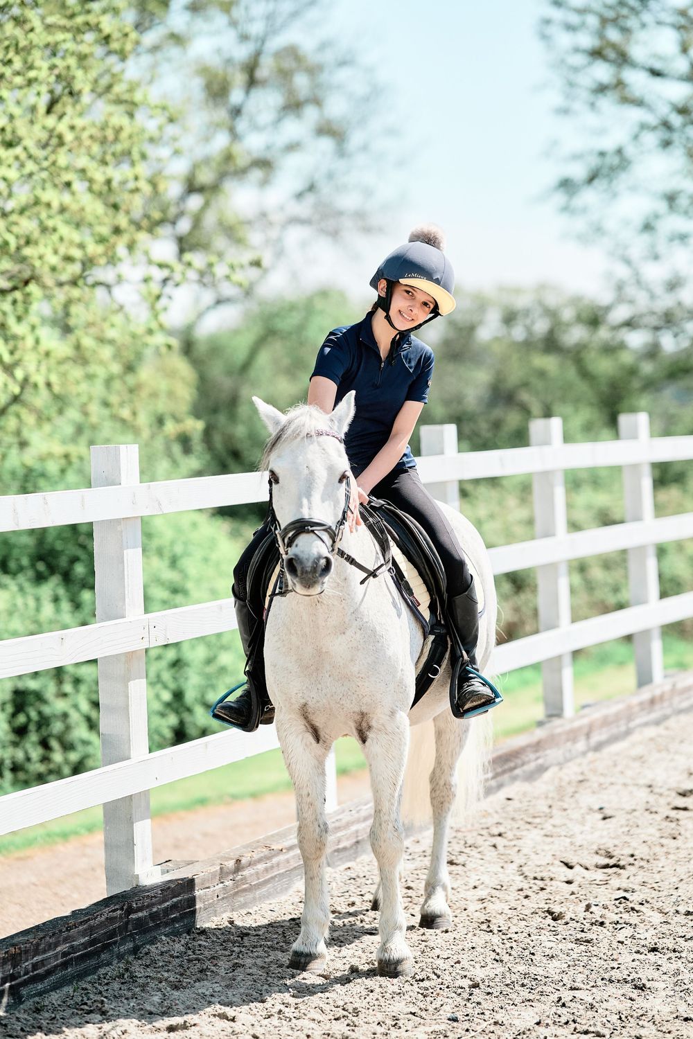 A rider in black attire practices horseback riding on a white horse along a white fence in an outdoor arena.