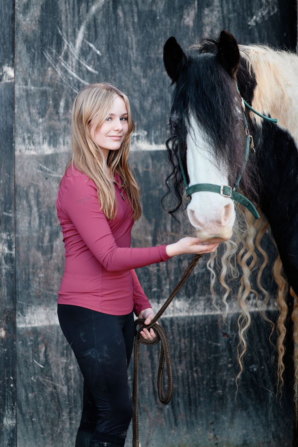 Woman in burgundy sweater grooms and brushes a black horse's long mane against dark textured wall.