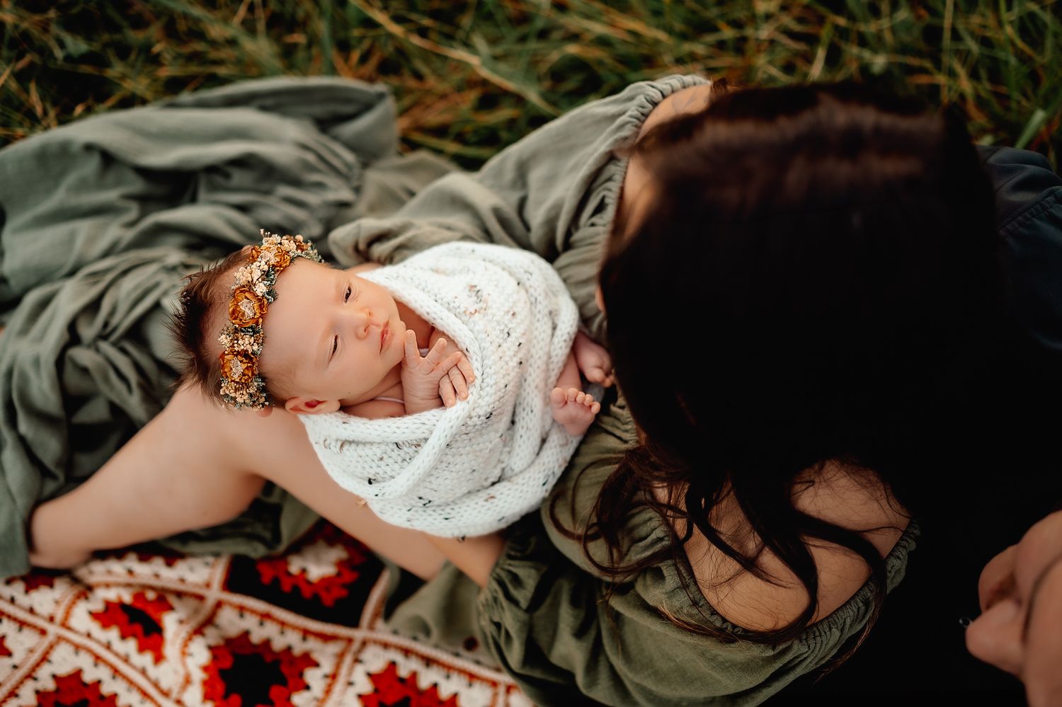 A sleeping baby wrapped in white blanket on a patterned blanket outdoors.