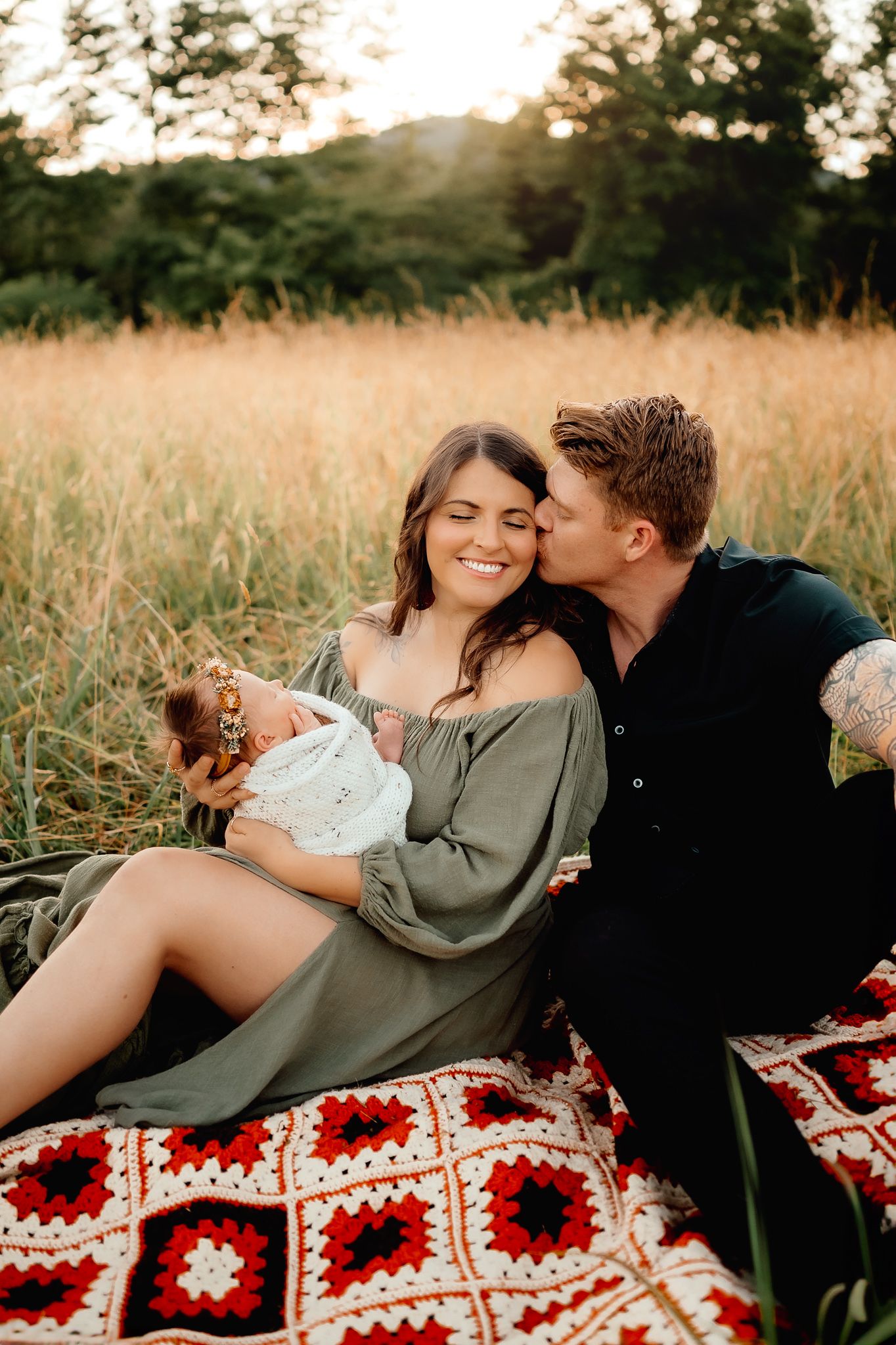 Family relaxing on a red patterned blanket in a sunlit meadow.