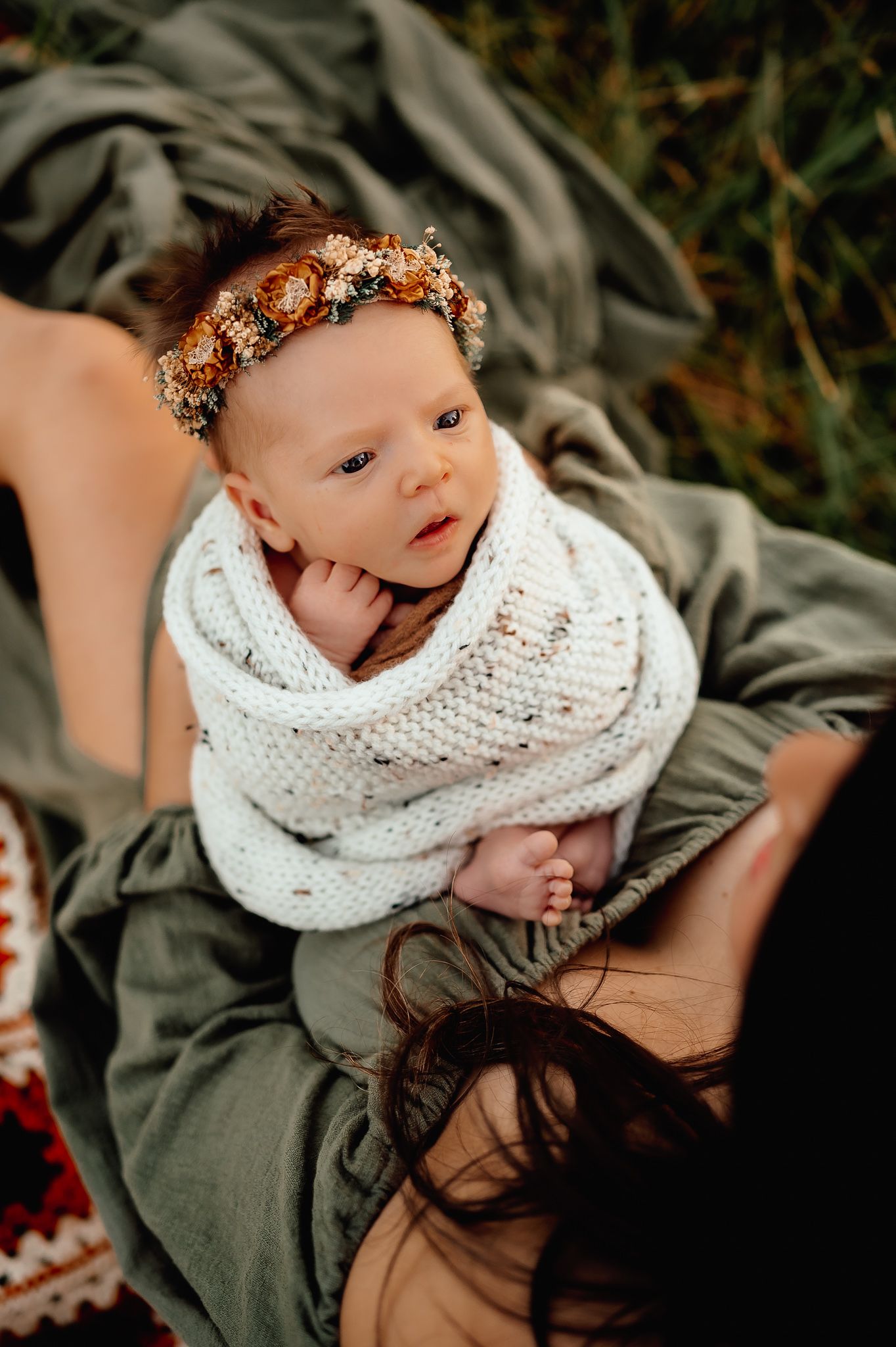 Baby wearing floral crown and white knit wrap looking curiously at camera.