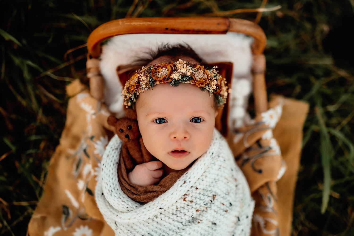 Baby with floral crown wrapped in white knit blanket lying in rustic basket.