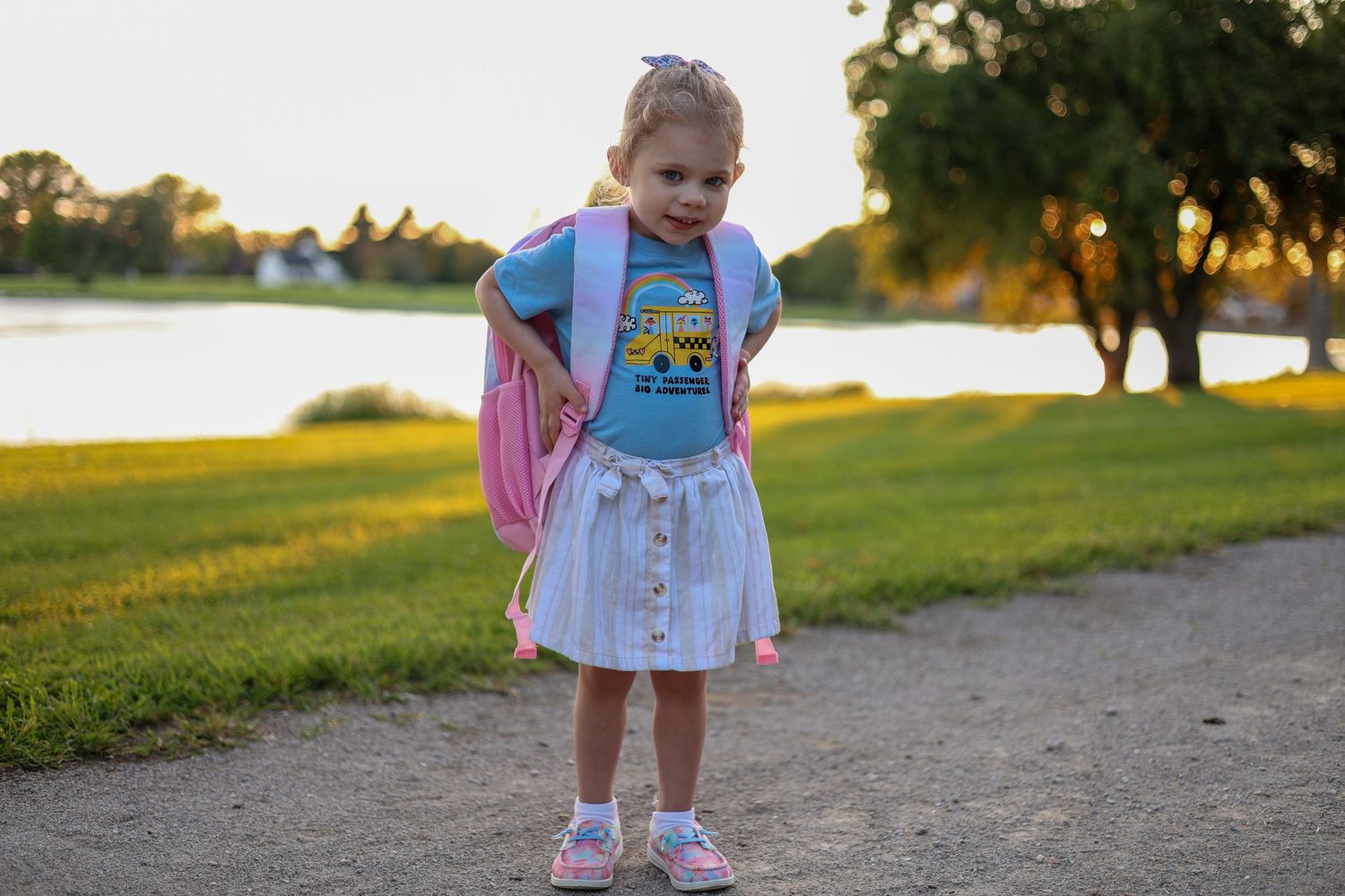 Child stands on path wearing Minions shirt and pink backpack with lake in background.