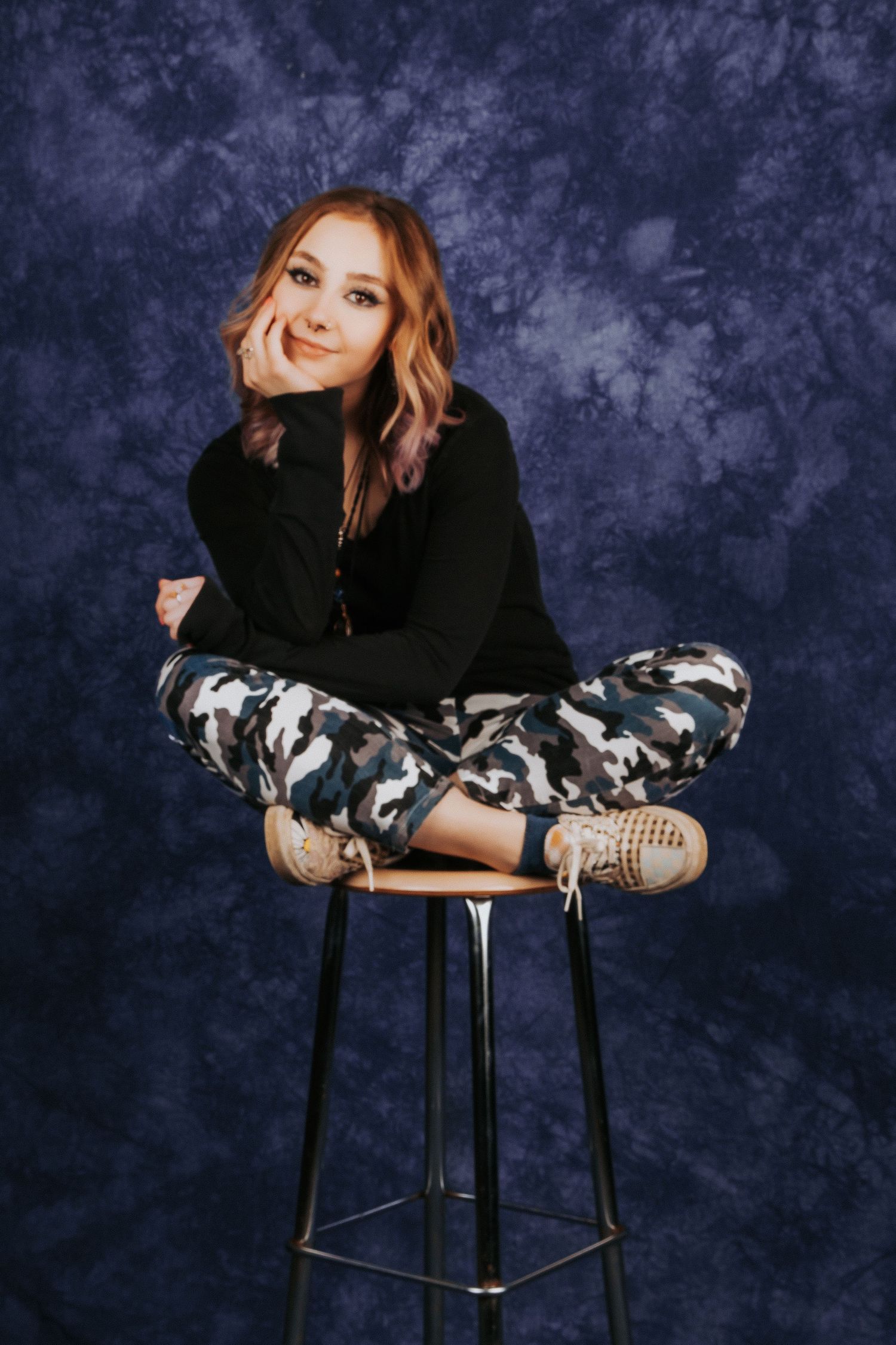 Model in black sweater and camo pants poses playfully on barstool against textured background.