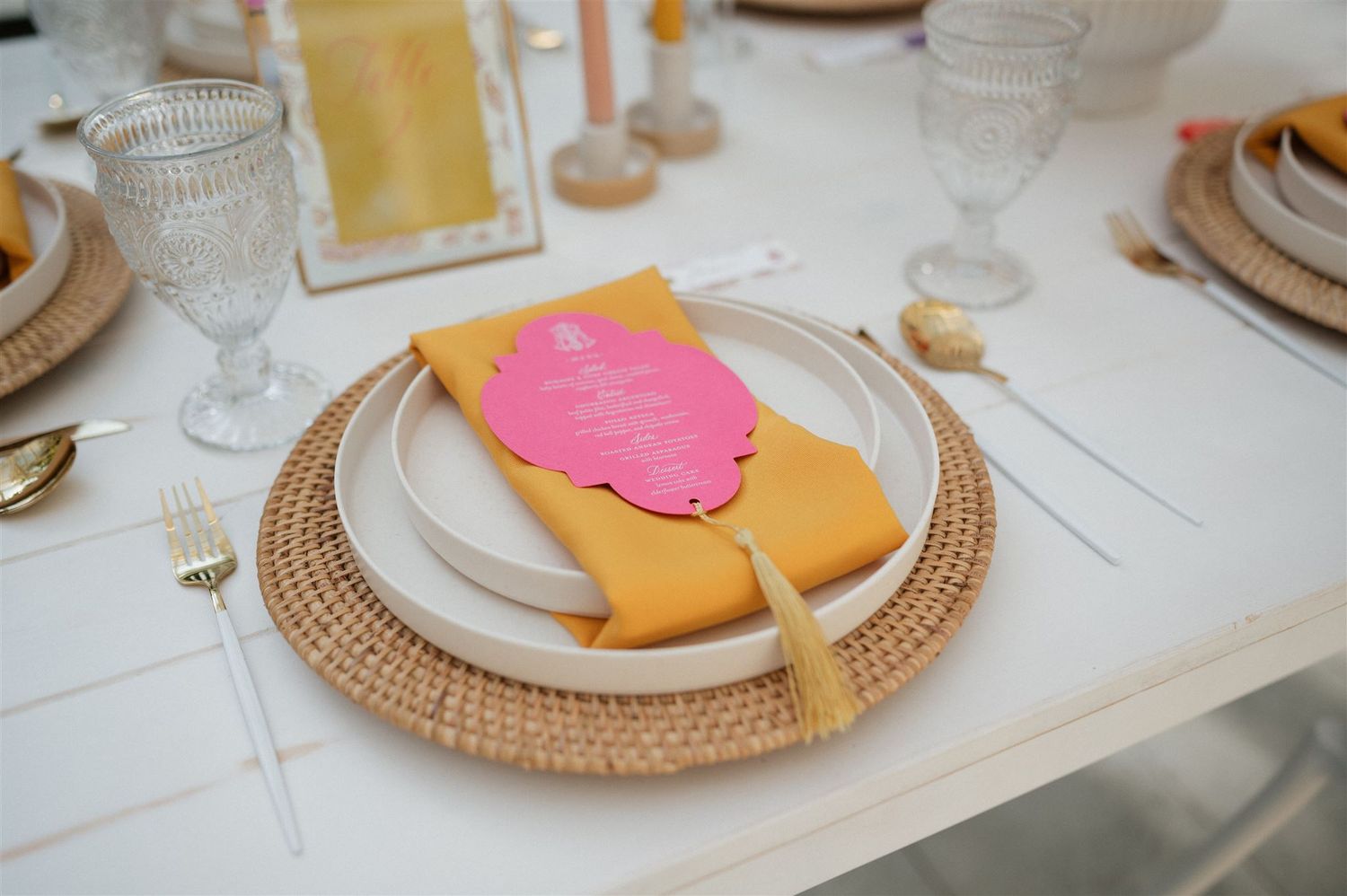 Table place setting with white plates on rattan charger featuring pink and yellow napkins with gold cutlery.